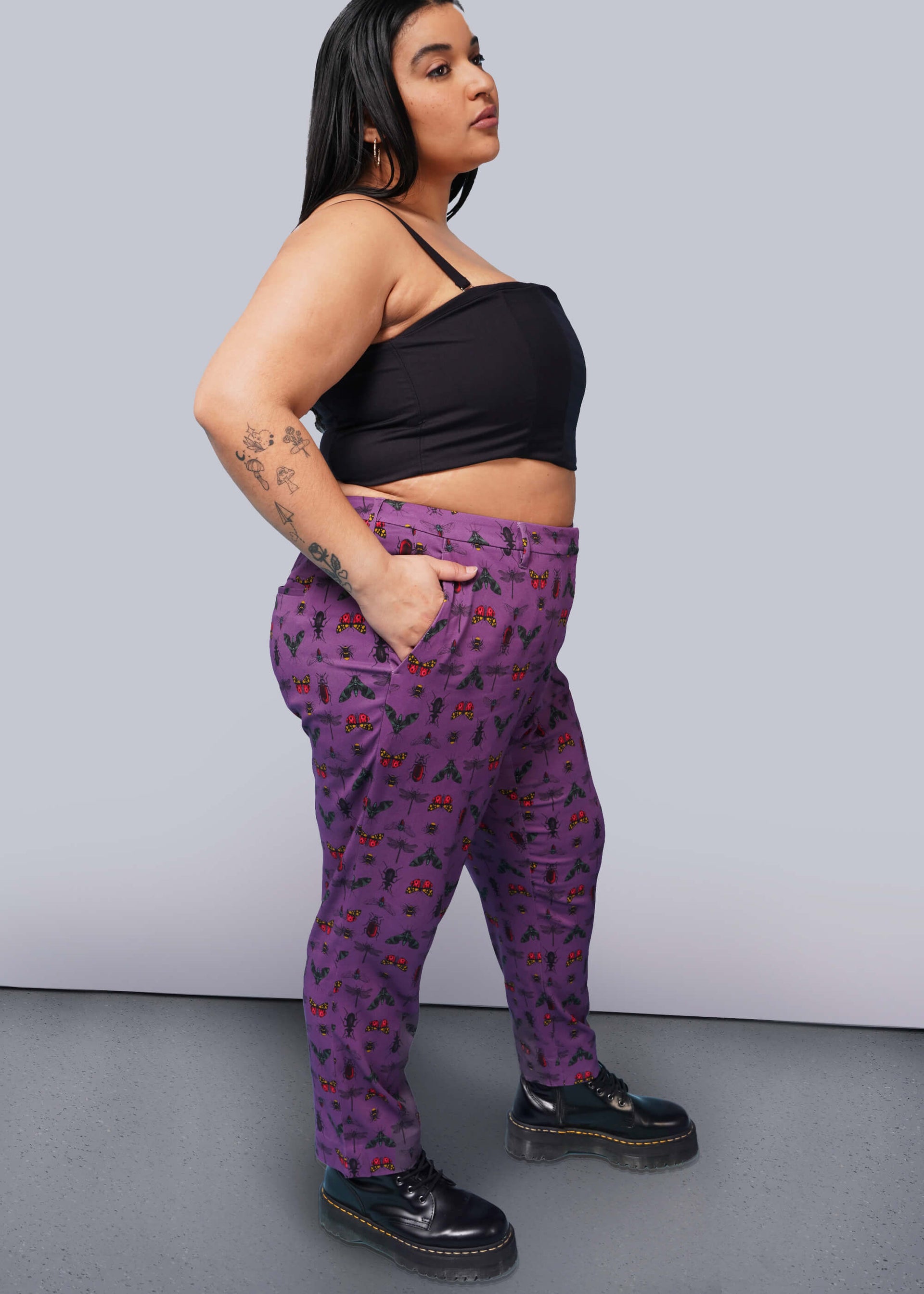 1X=Side view of person with long black hair with hand in pocket wearing black corset, purple bug printed slim crop pants, and Doc Marten boots.
