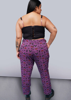 1X= Back view of person with long black hair with hand in pocket wearing black corset, purple bug printed slim crop pants, and Doc Marten boots.