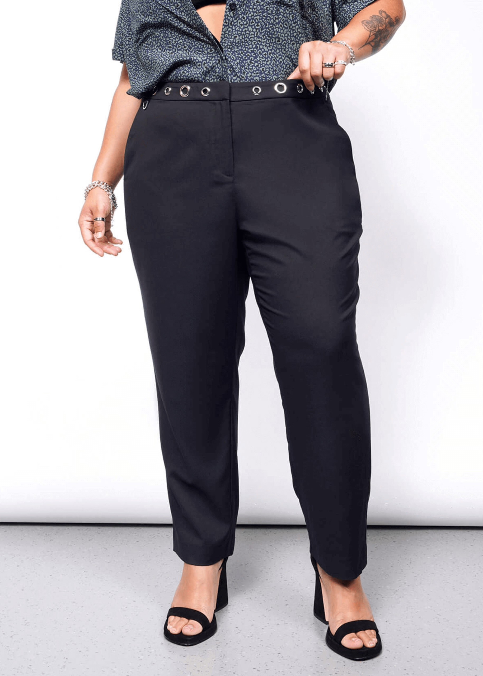 1X= A tattooed person on their left forearm wears Debbie Harry X Wildfang Slim Crop Pant in black, highlighted by large grommets, paired with a punk-style patterned blue shirt. They stand on a gray floor in black heeled sandals, with their right hand resting on the waistband.