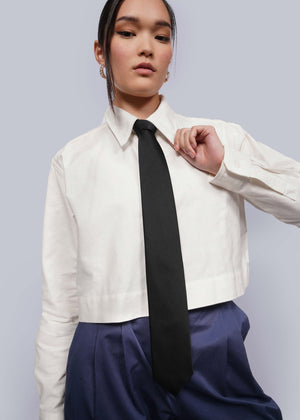 Person with black hair tied back wears cropped white button up, navy pants, and black silk tie.