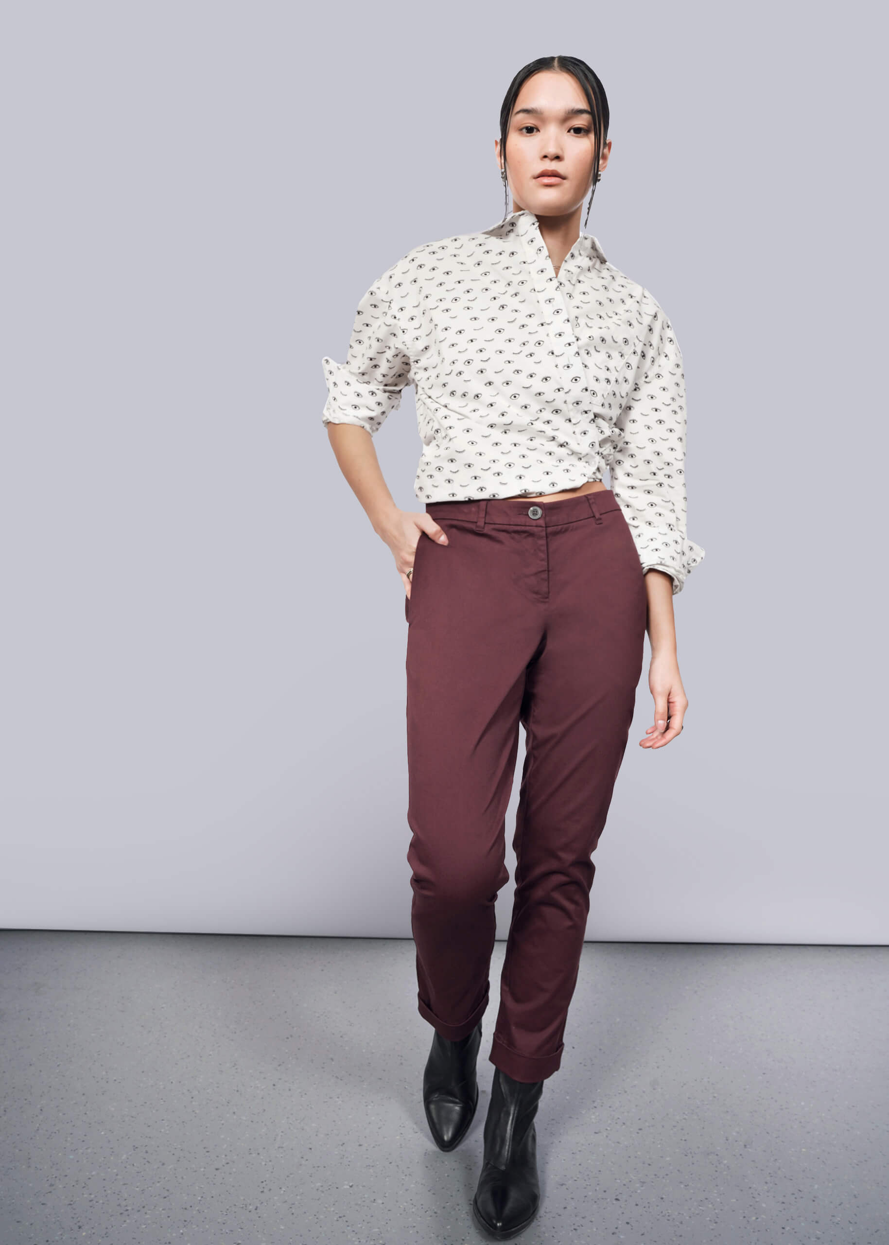 S=A person stands against a white backdrop wearing a patterned white shirt with convertible cuffs, The Essential Trouser in burgundy tapered-leg style, and black ankle boots. One hand is casually in a pocket, their expression neutral.