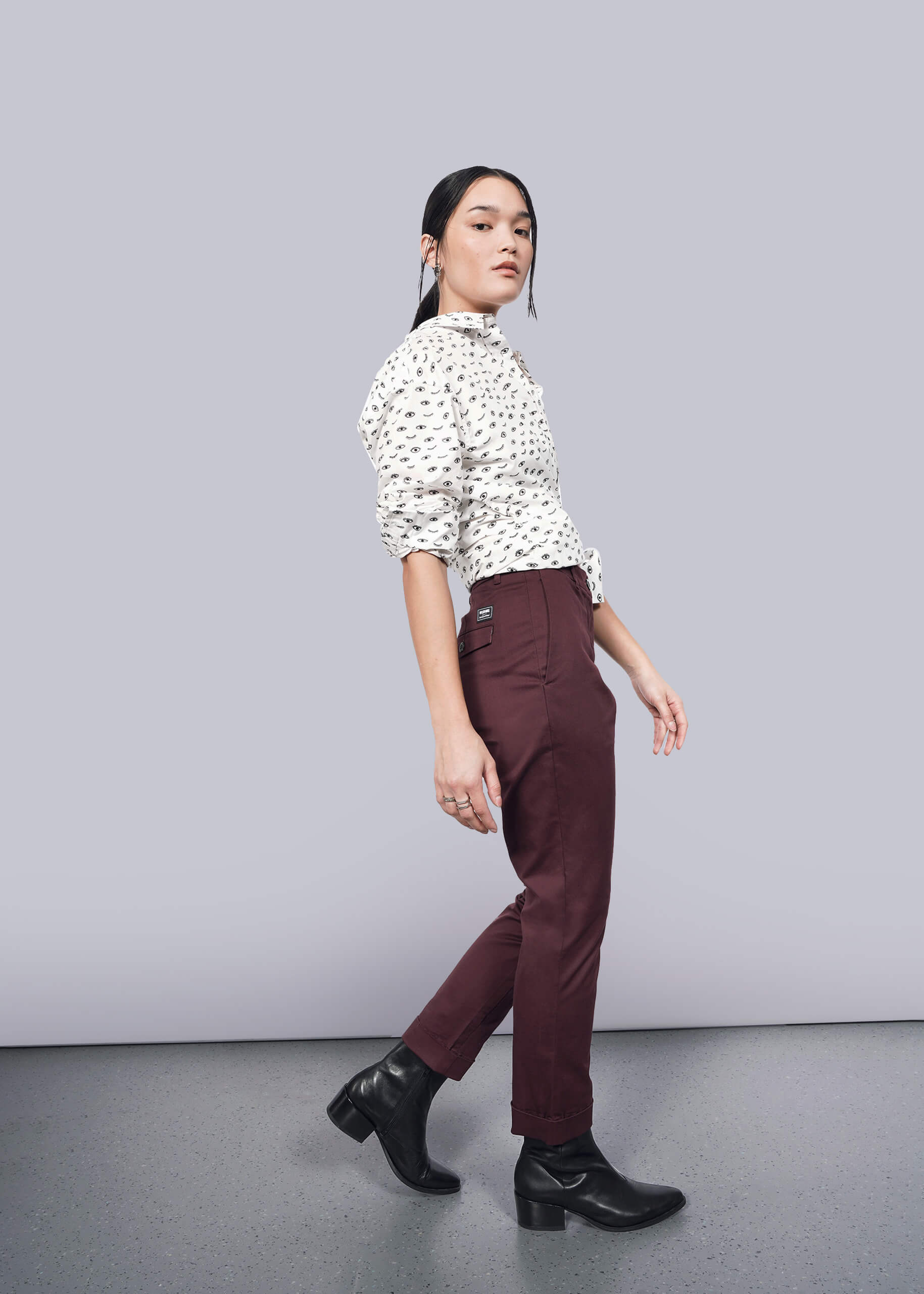 S=A person wearing a white patterned blouse with The Essential Trouser and black ankle boots poses sideways against a plain white background, gazing forward.