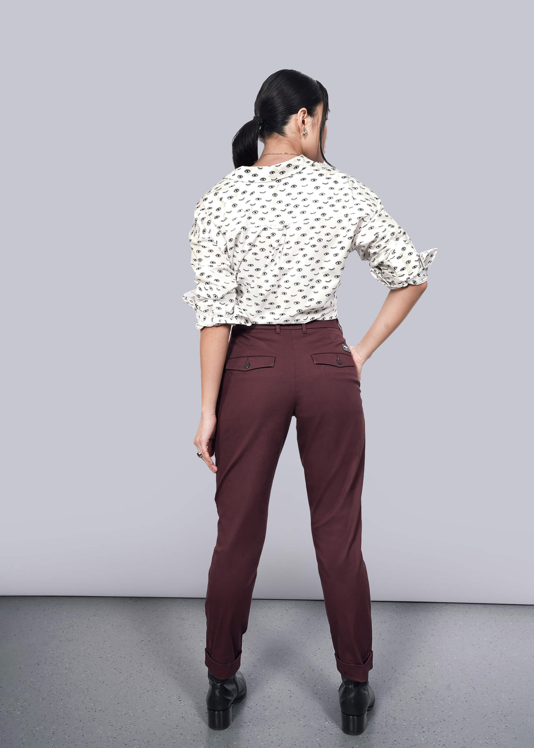 S=A woman stands facing away in The Essential Trouser, maroon with a tapered leg, paired with a white blouse featuring a black pattern. Her hair is tied back, and shes set against a plain white background on a speckled gray floor.