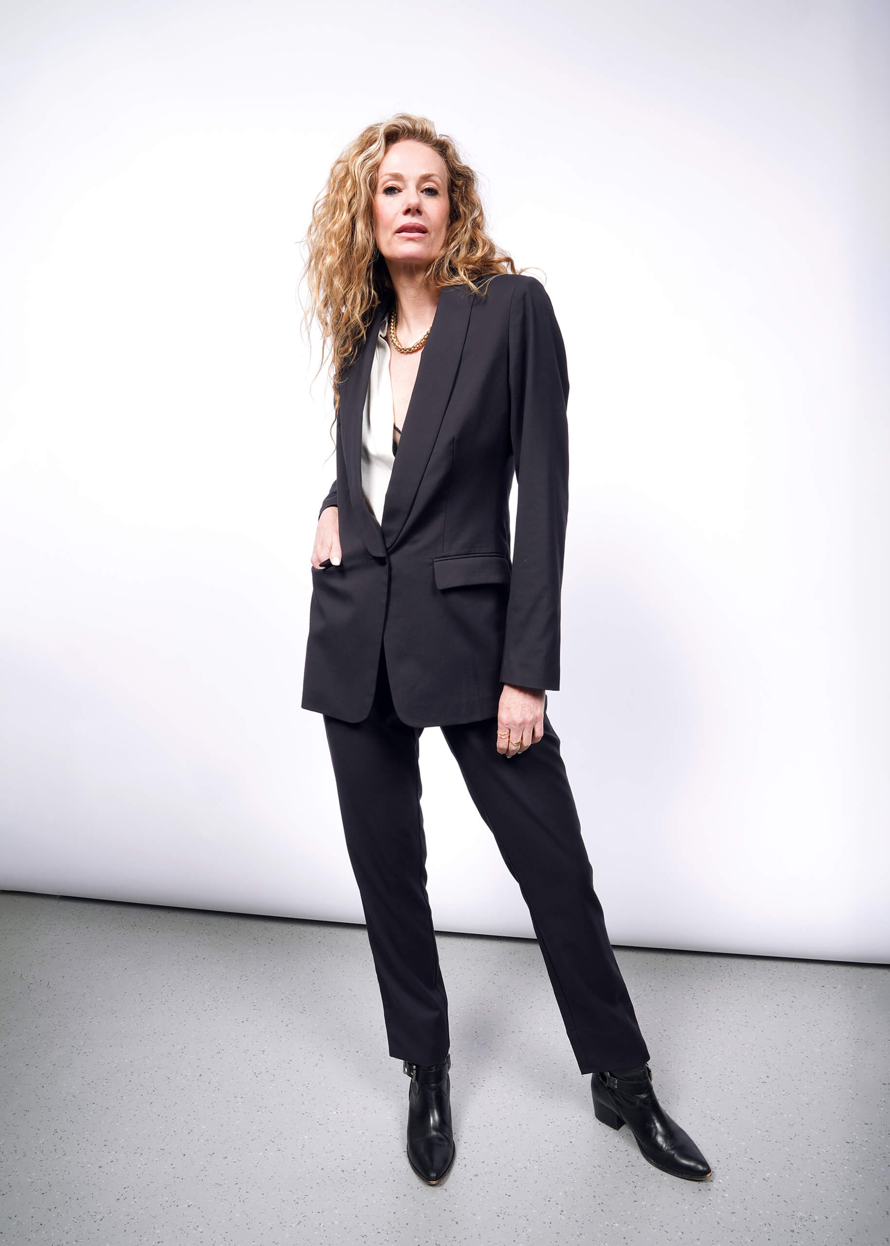 S=A person with long curly hair confidently stands against a plain white background in The Empower Tux Blazer, a black suit, and boots. Their left hand rests in the deep pocket of their pants, showcasing a relaxed yet assertive pose that complements the blazers timeless tailored fit.
