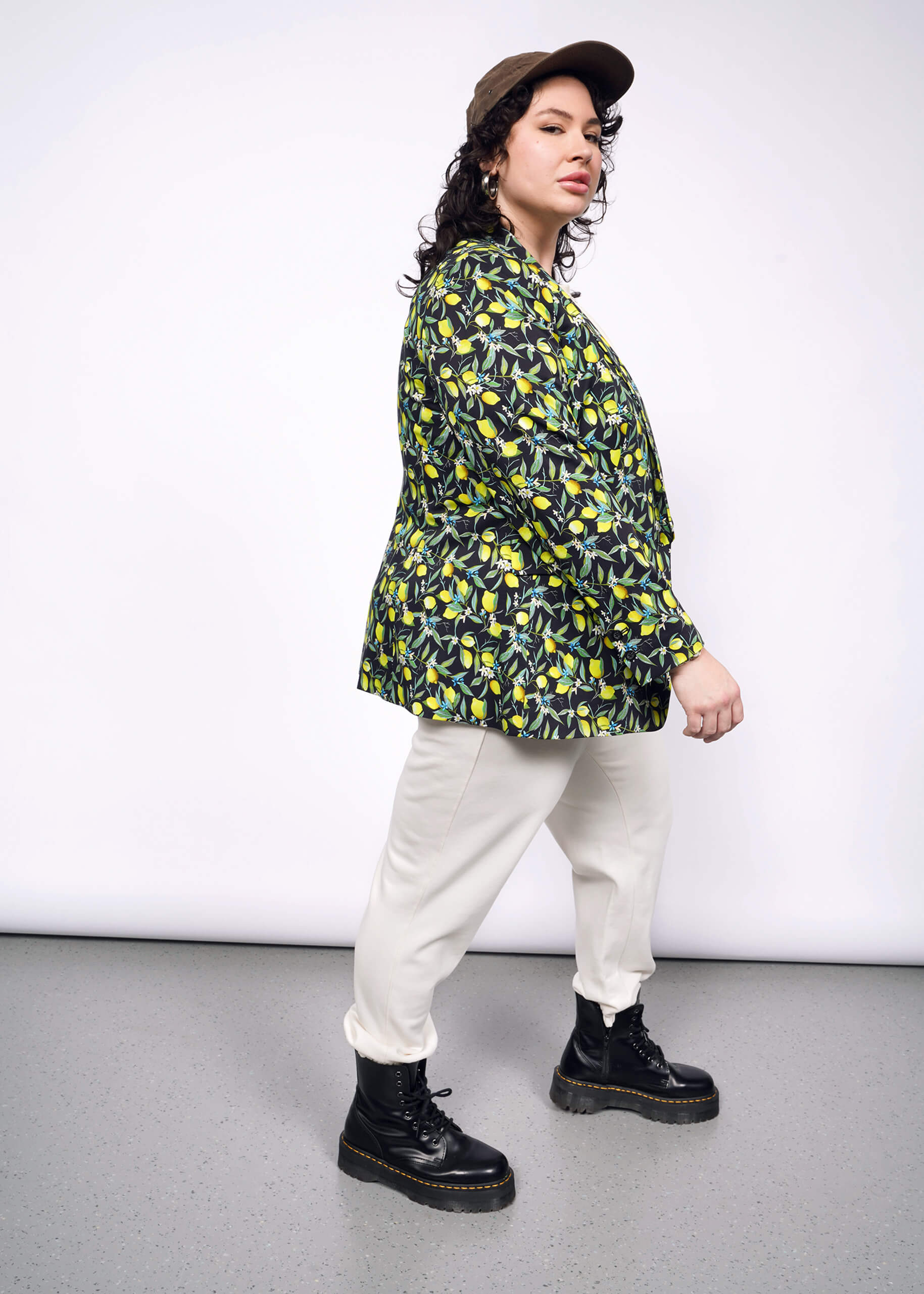 1X=Side full body view of person with short curly black hair wearing black cap, tan colored shirt and pants, black boots, and The Empower Tux Blazer in Citrus Black, which is a yellow and green print on black background featuring lemons and leaves.