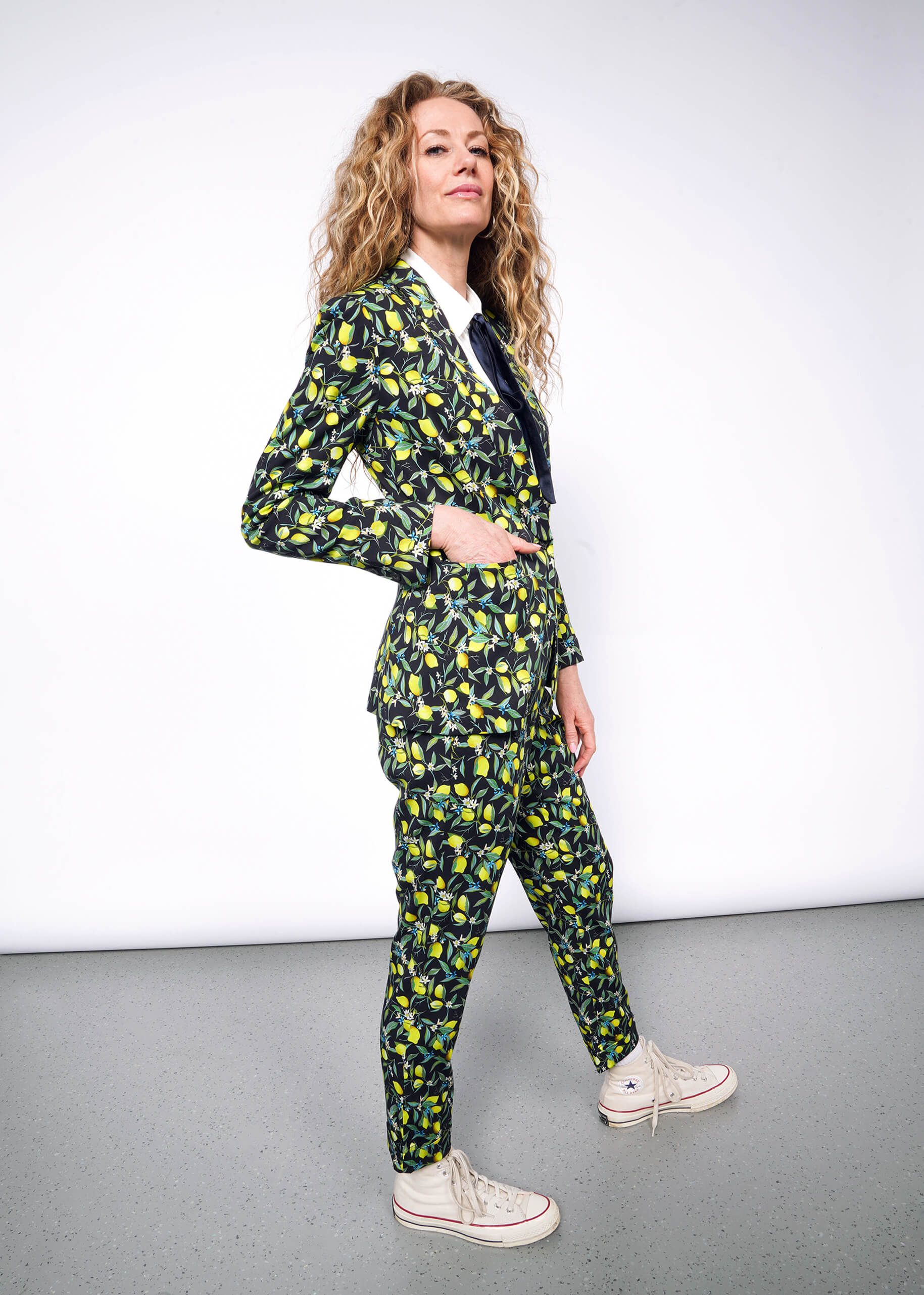 S= Side full body view of person with curly blonde hair wearing The Empower Bow Button Up in white, with The Empower Tux Blazer and The Empower Trouser in Citrus Black, which is a yellow and green print on black background featuring lemons and leaves. They also wear white Converse sneakers and have their hand in the blazer pocket.