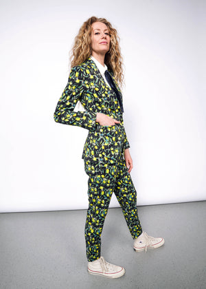 S= Side full body view of person with curly blonde hair wearing The Empower Bow Button Up in white, with The Empower Tux Blazer and The Empower Trouser in Citrus Black, which is a yellow and green print on black background featuring lemons and leaves. They also wear white Converse sneakers and have their hand in the blazer pocket.