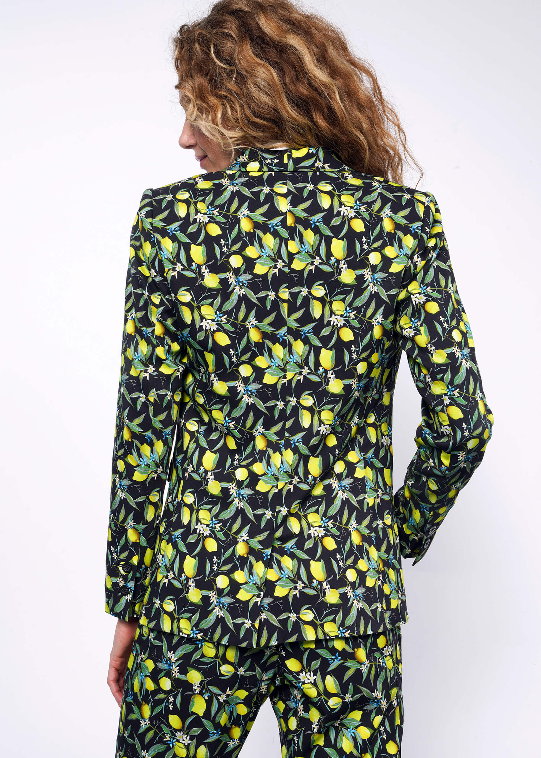 S=Back view of person with curly blonde hair wearing The Empower Tux Blazer and The Empower Trouser in Citrus Black, which is a yellow and green print on black background featuring lemons and leaves.