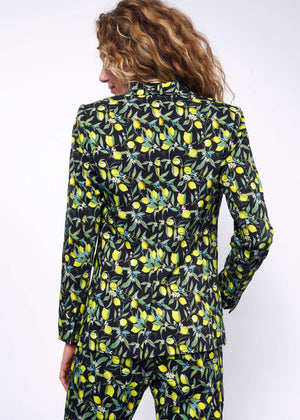 S=Back view of person with curly blonde hair wearing The Empower Tux Blazer and The Empower Trouser in Citrus Black, which is a yellow and green print on black background featuring lemons and leaves.