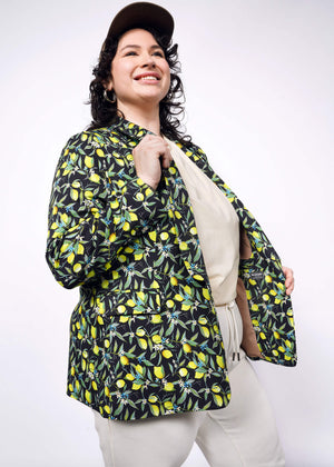 1X=Person with short curly black hair wear black cap, tan colored shirt and pants, with The Empower Tux Blazer in Citrus Black, which is a yellow and green print on black background featuring lemons and leaves.