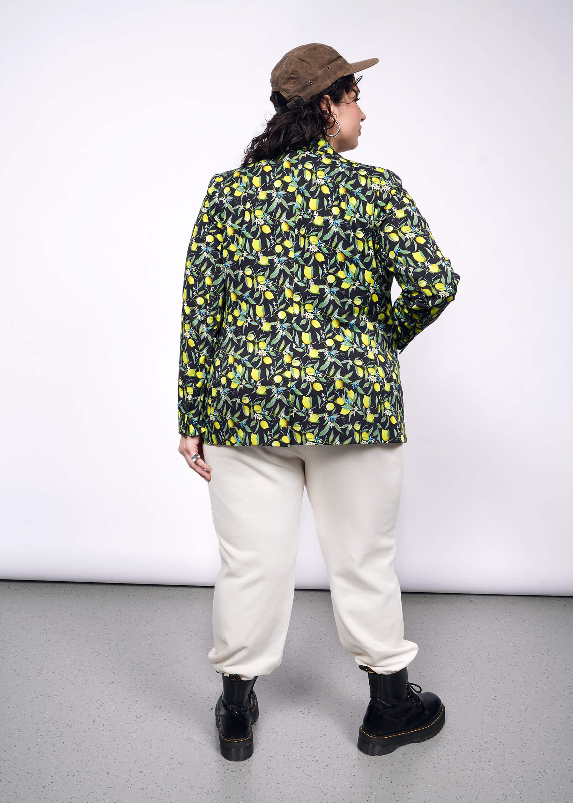 1X=Back view of person with short curly black hair wearing black cap, tan colored pants, black boots, and The Empower Tux Blazer in Citrus Black, which is a yellow and green print on black background featuring lemons and leaves.