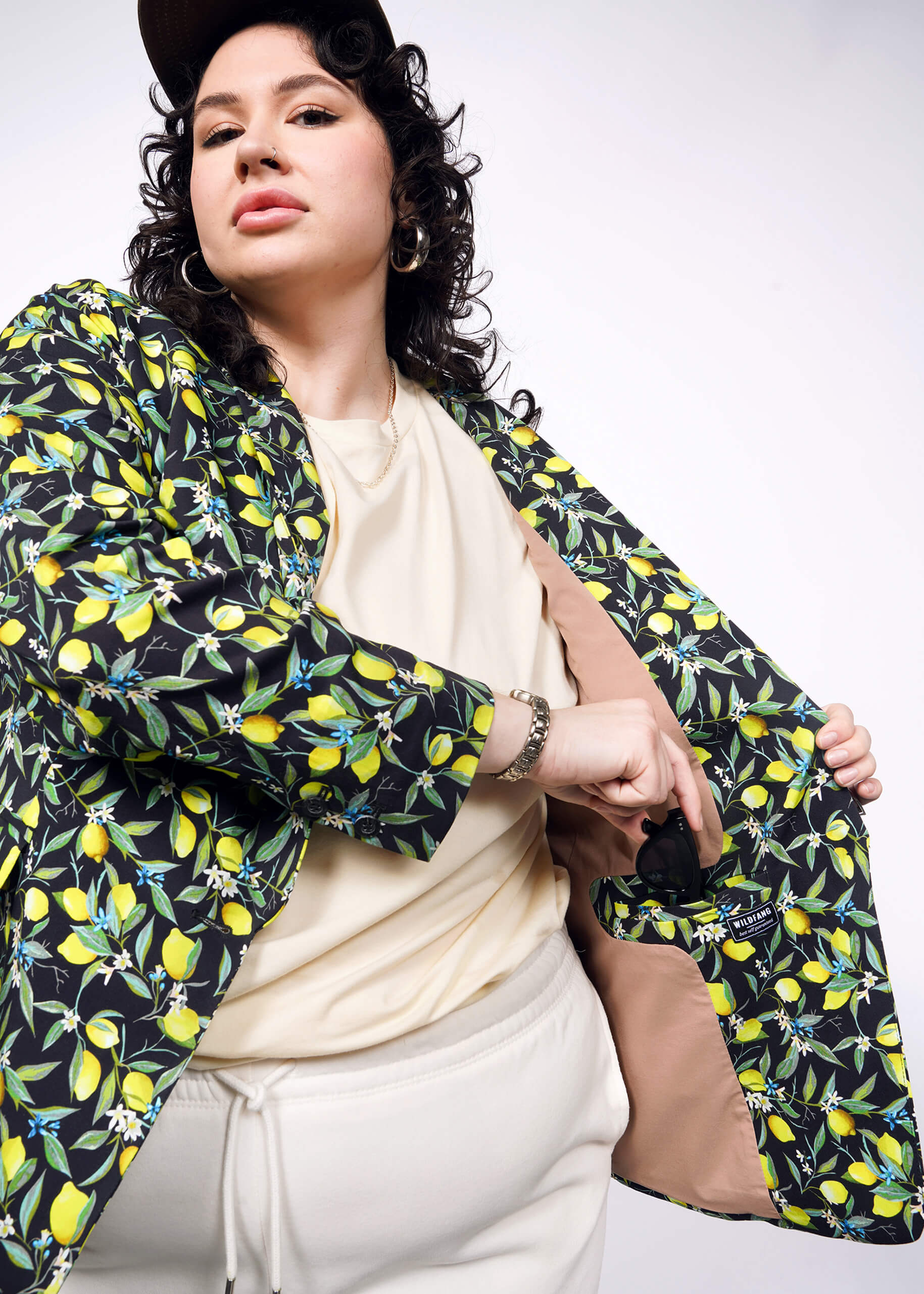 1X=Person with short curly black hair wear black cap, tan colored shirt and pants, with The Empower Tux Blazer in Citrus Black, which is a yellow and green print on black background featuring lemons and leaves. They hold blazer open and are putting sunglasses in the interior blazer pocket.