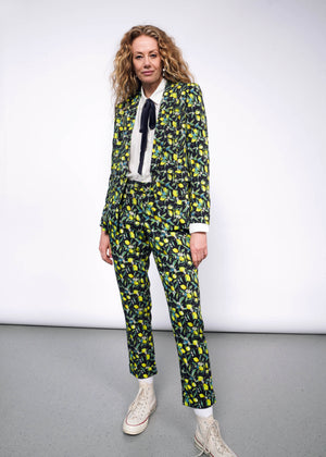 S=Front full body view of person with curly blonde hair wearing The Empower Bow Button Up in white, with The Empower Tux Blazer and The Empower Trouser in Citrus Black, which is a yellow and green print on black background featuring lemons and leaves. They also wear white Converse sneakers.