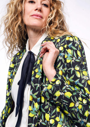 S=Close up side view of person with curly blonde hair wearing The Empower Bow Button Up in white, with The Empower Tux Blazer in Citrus Black, which is a yellow and green print on black background featuring lemons and leaves.