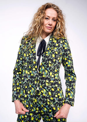 S= Front view of person with curly blonde hair wearing The Empower Bow Button Up in white, with The Empower Tux Blazer and The Empower Trouser in Citrus Black, which is a yellow and green print on black background featuring lemons and leaves.