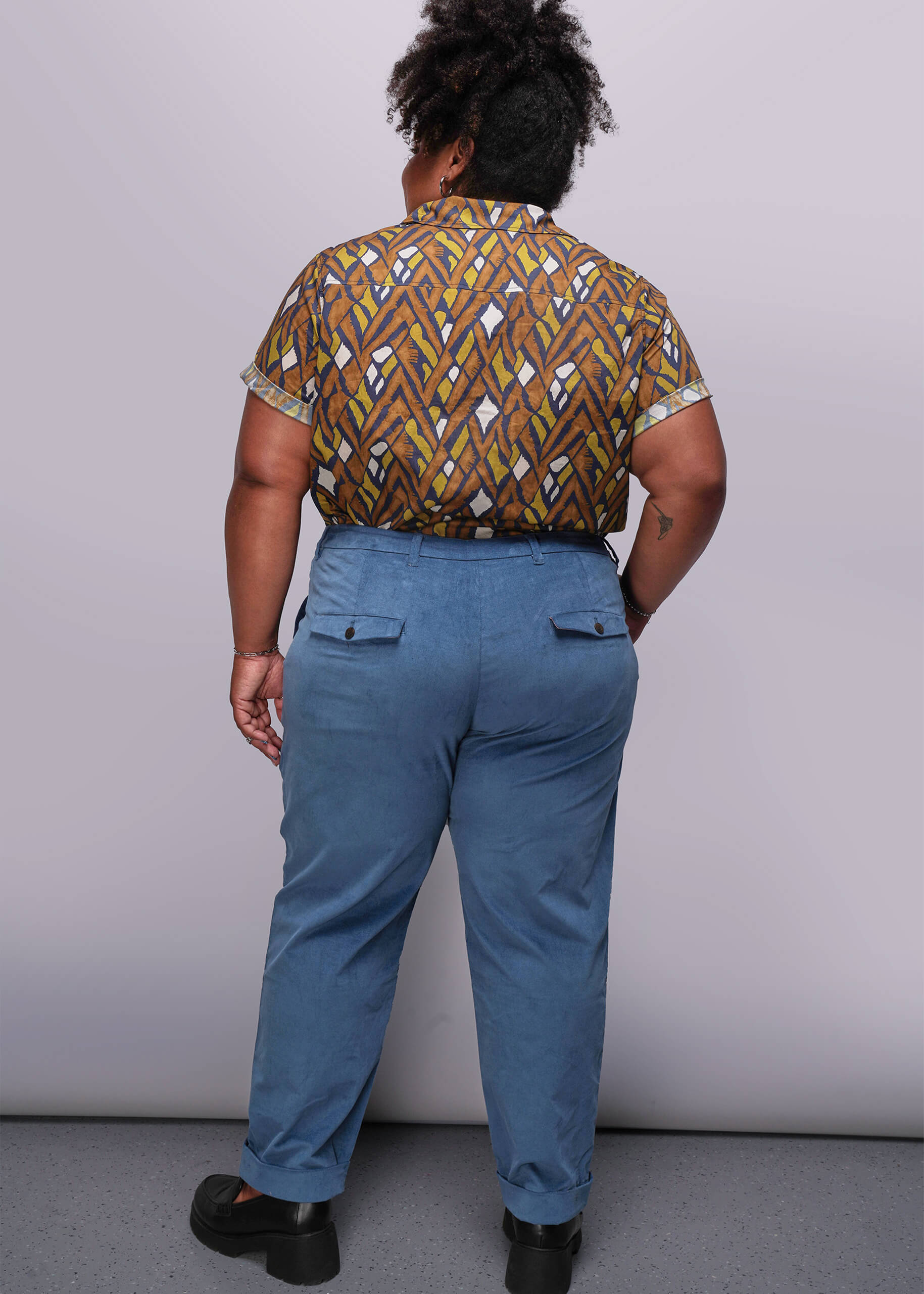 1X=Person wearing a patterned shirt and blue pants against a plain background