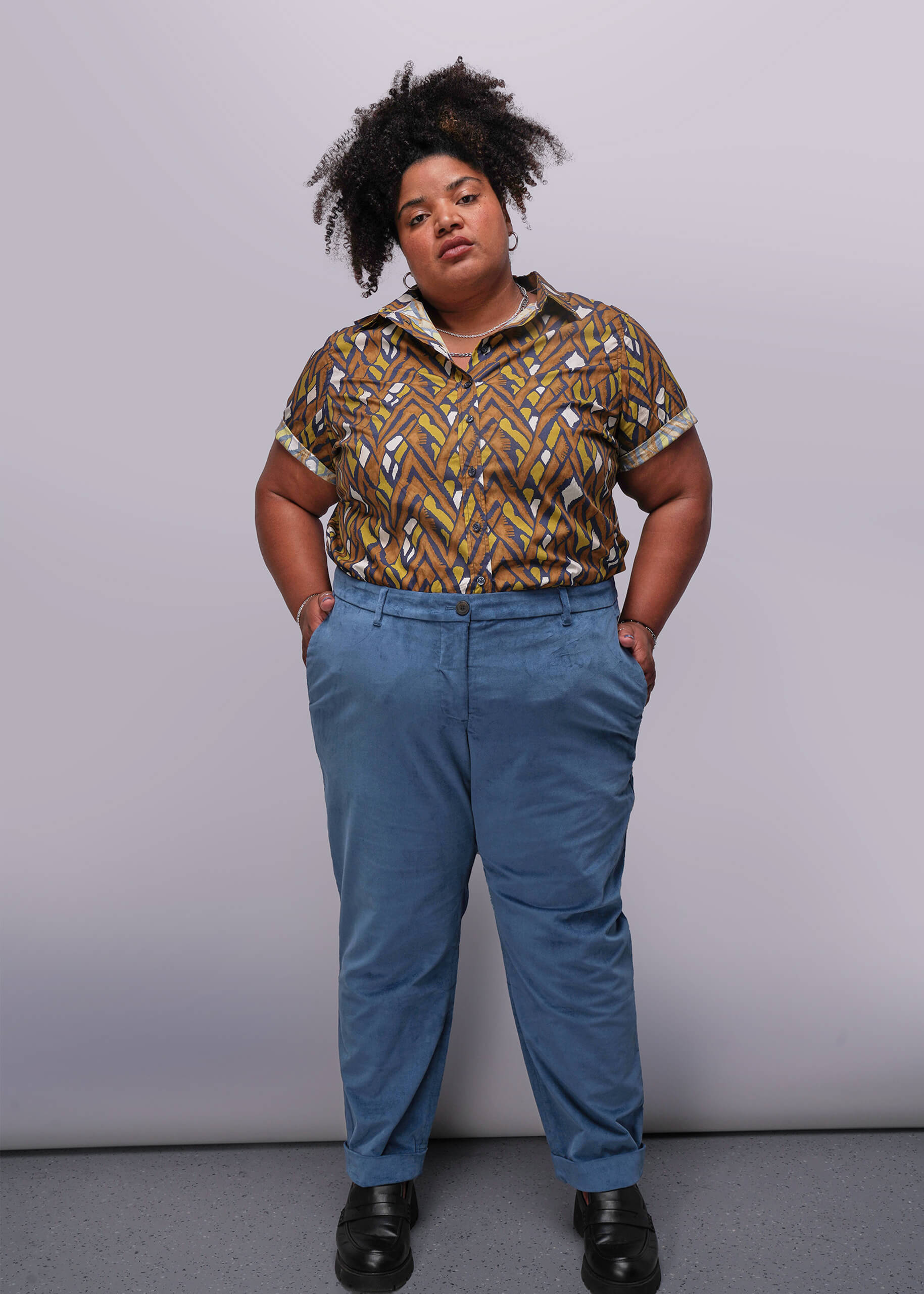 1X=Person wearing a patterned shirt and blue pants against a plain background