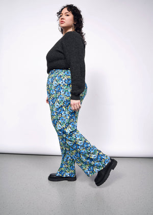 1X=Side view of person with curly short black hair looking at camera wearing black sweater tucked into patterned floral wide leg trousers and black boots.