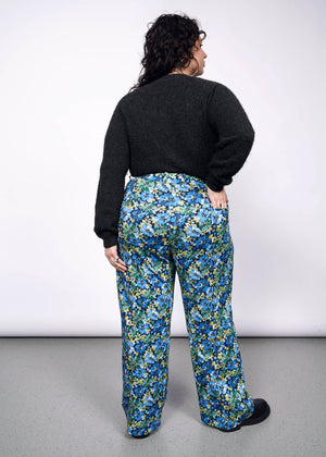 1X=Back view of person with curly short black hair wearing black sweater tucked into patterned floral wide leg trousers and black boots.
