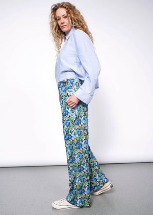 S=Side view of person with medium length wavy blonde hair looking at camera wearing light blue button up shirt tucked into patterned floral blue and green wide leg trousers and Converse sneakers. Neutral background.