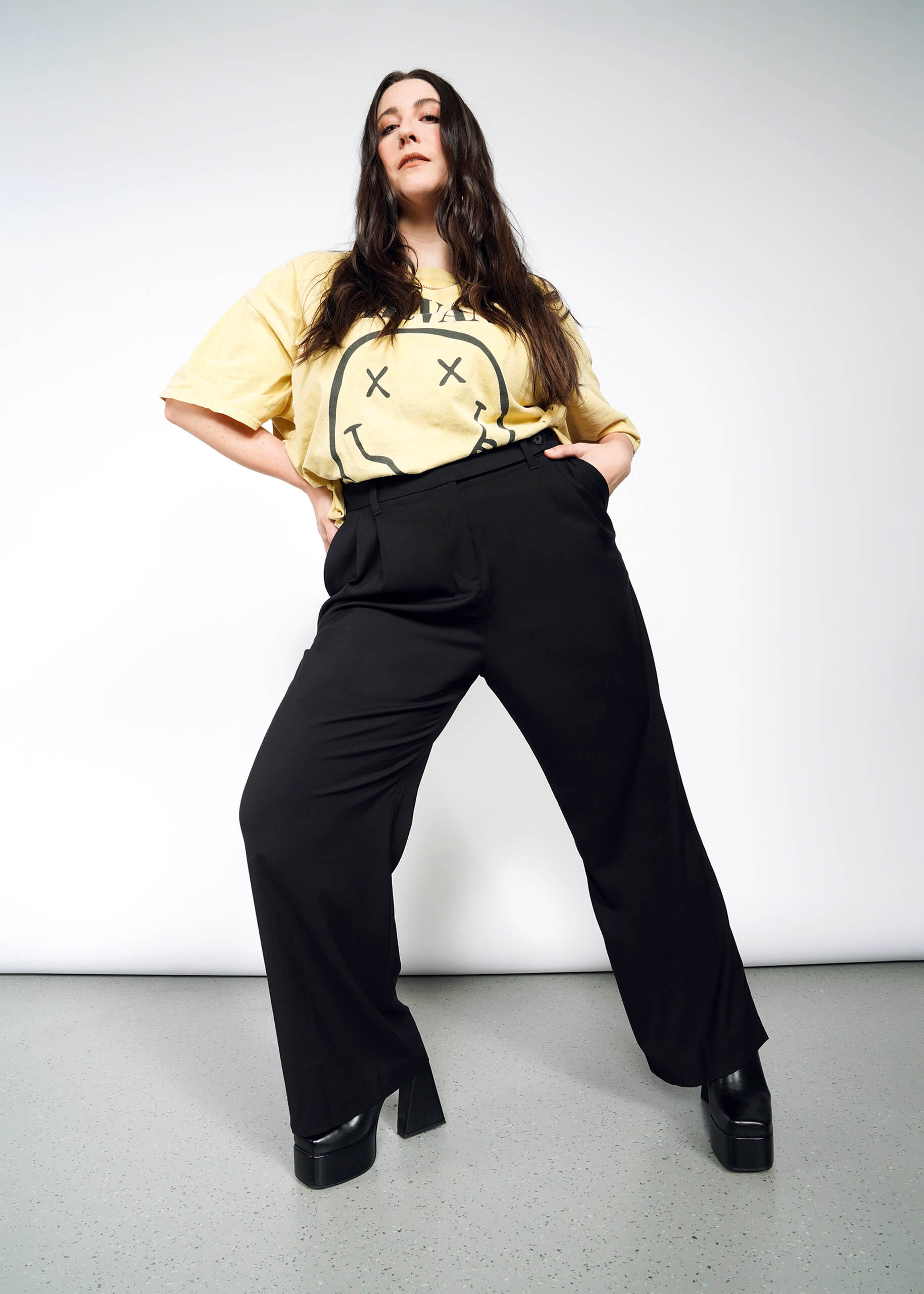 1X=A woman with long brown hair and a neutral expression stands confidently with hands in her pockets, wearing The Empower Wide Leg Trouser in black, paired with a yellow cartoon-face t-shirt and black platform shoes.