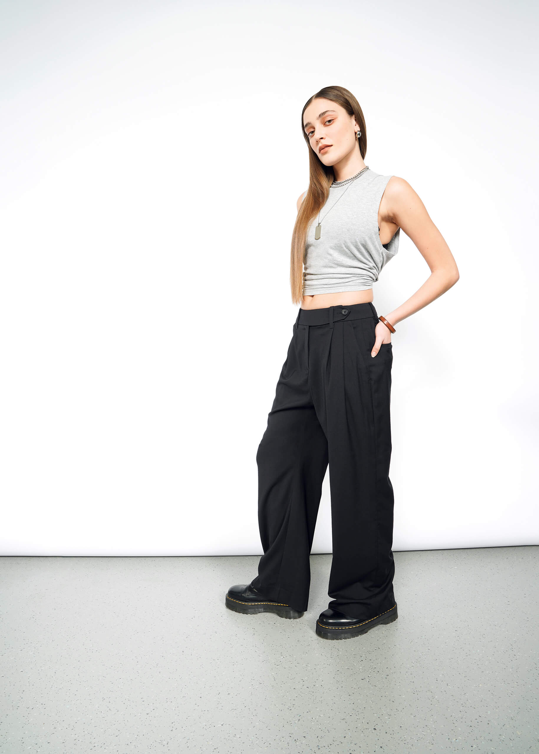 S=A woman with straight, long hair and a neutral expression stands against a white background, wearing The Empower Wide Leg Trouser with a sleeveless grey crop top, black platform shoes, and layered necklaces.