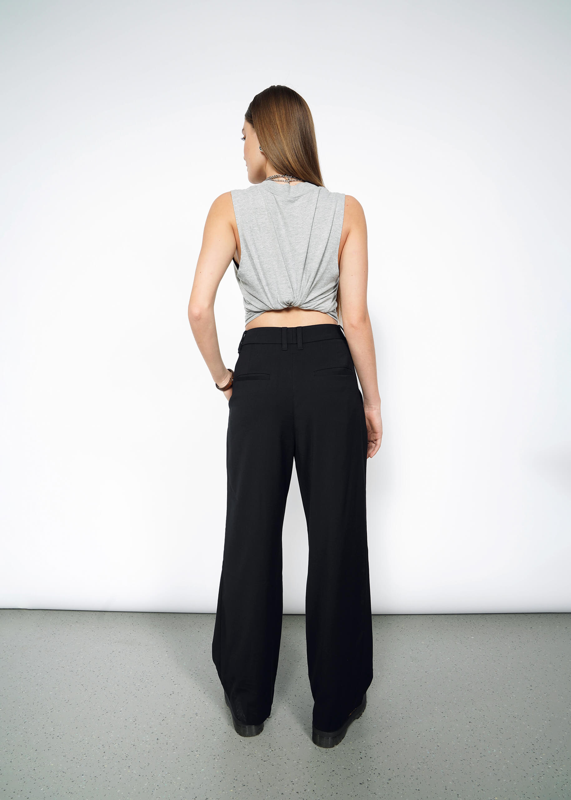 S=A person with long brown hair stands with their back to the camera, wearing a sleeveless gray top in a knot and The Empower Wide Leg Trouser for a stylish, modern look.