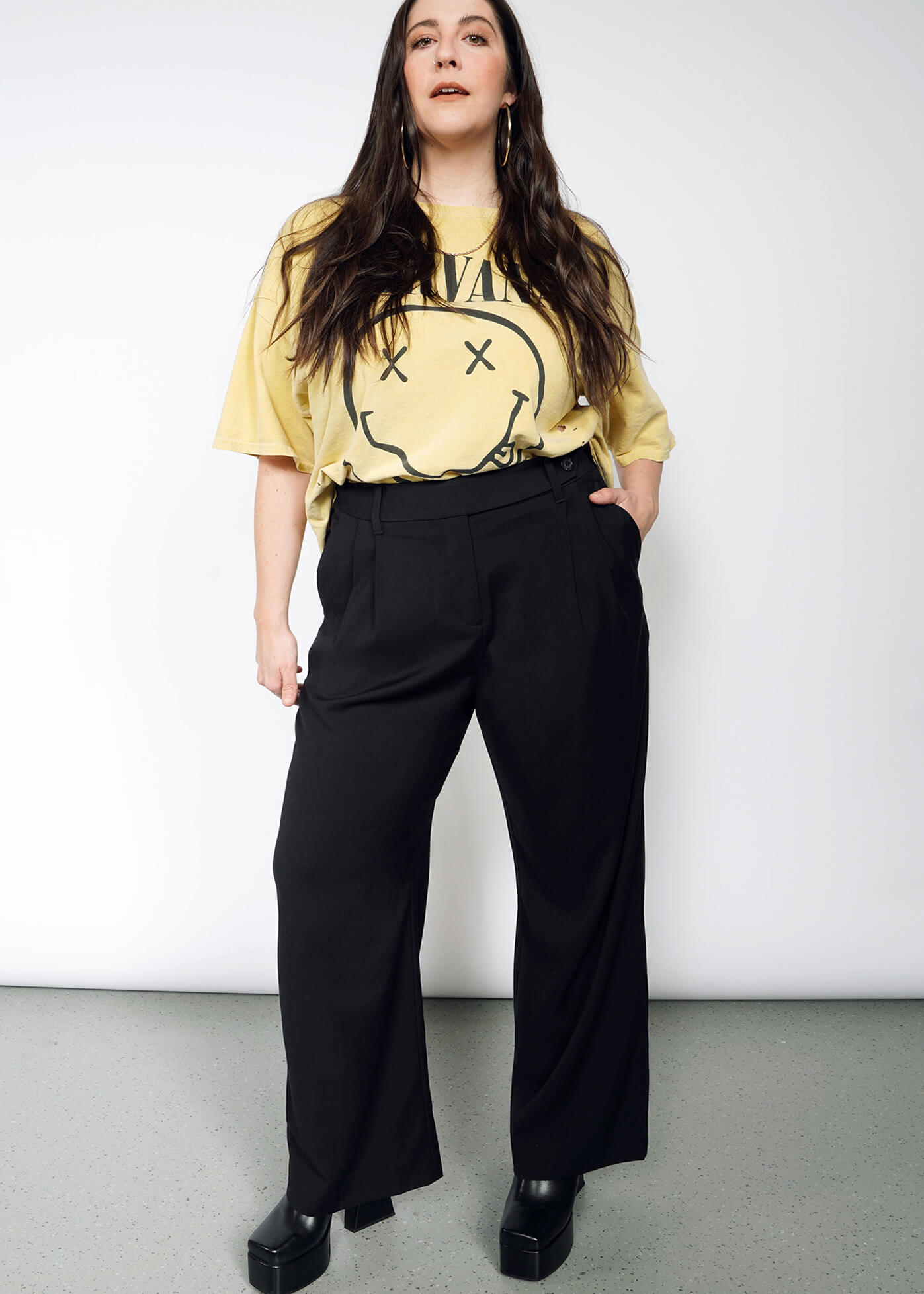 1X=Woman with long brown hair stands against a white background, wearing a yellow Nirvana T-shirt, The Empower Wide Leg Trouser, and black platform shoes, with her hands in her pockets.