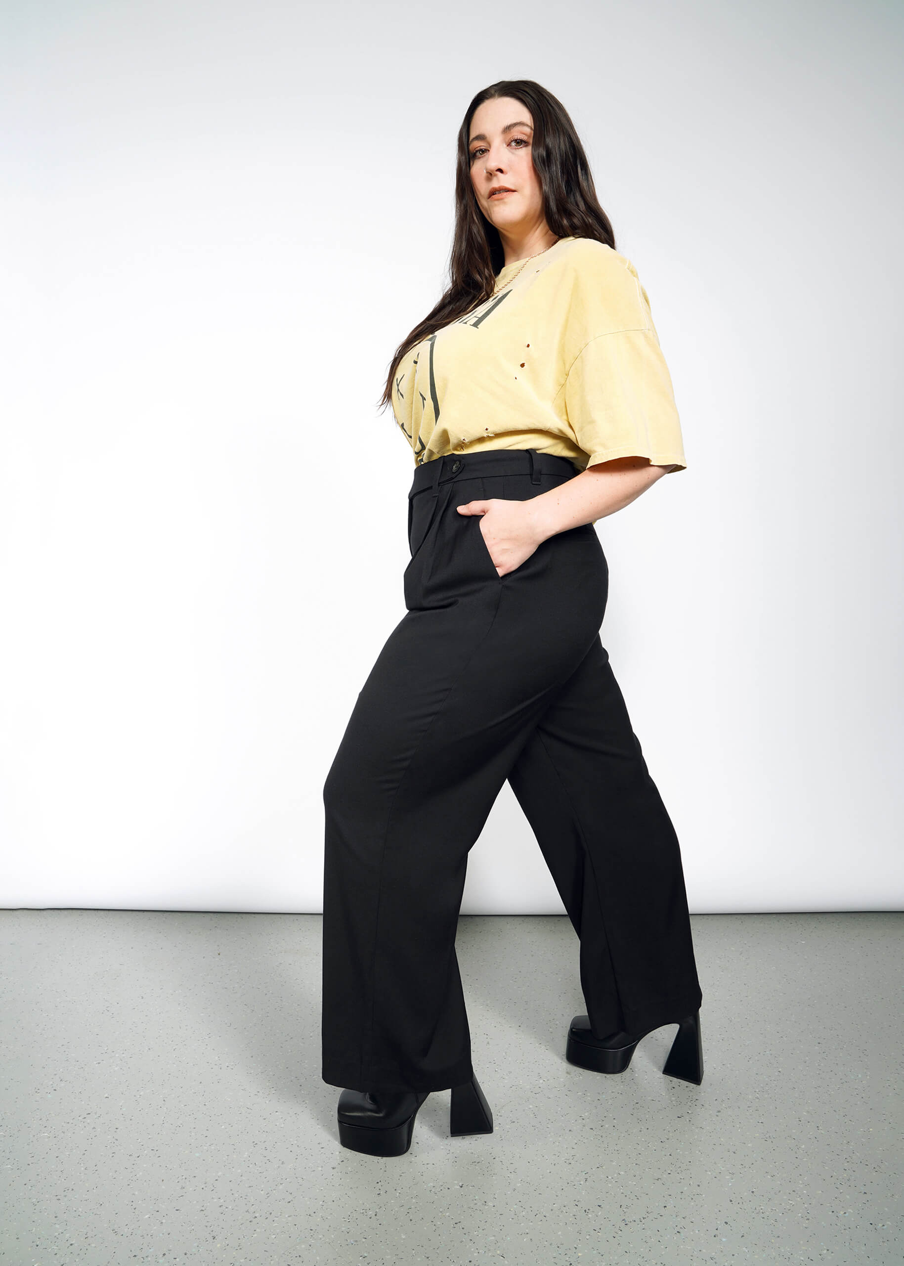 1X=A woman with long dark hair poses confidently in The Empower Wide Leg Trouser, a loose yellow tee tucked in, and black platform shoes against a plain light gray background.
