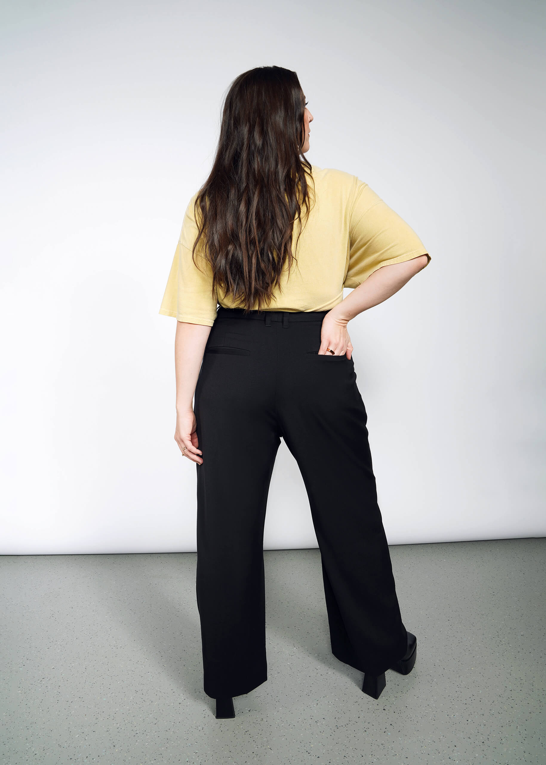 1X=A woman with long brown hair stands facing away, wearing a loose yellow shirt and The Empower Wide Leg Trouser—high-waisted, pleated-front pants with wide legs—one hand on her hip against a plain light background.