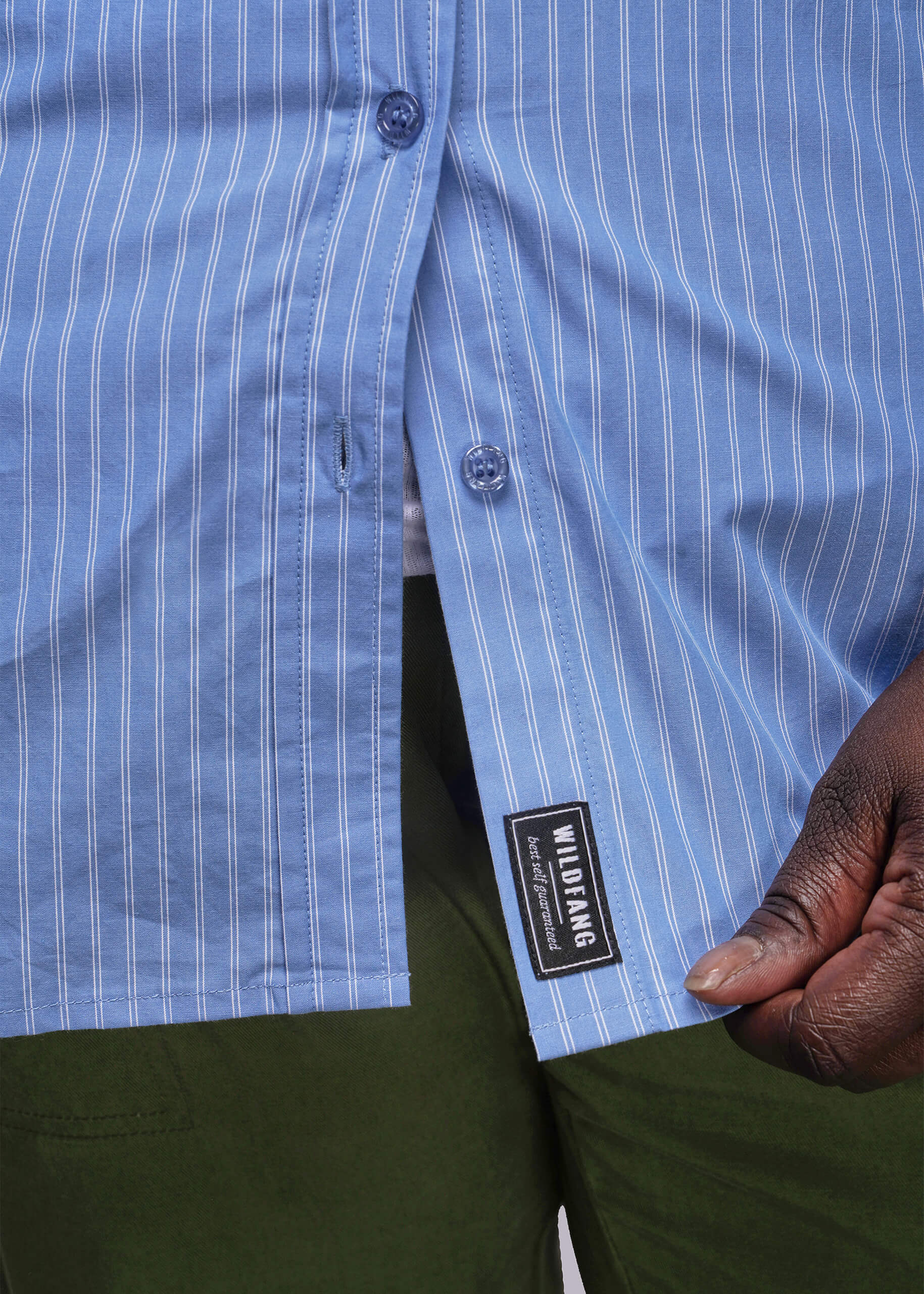 1X=Close-up of a blue striped shirt with a Wildfang label, held by a person wearing green pants.