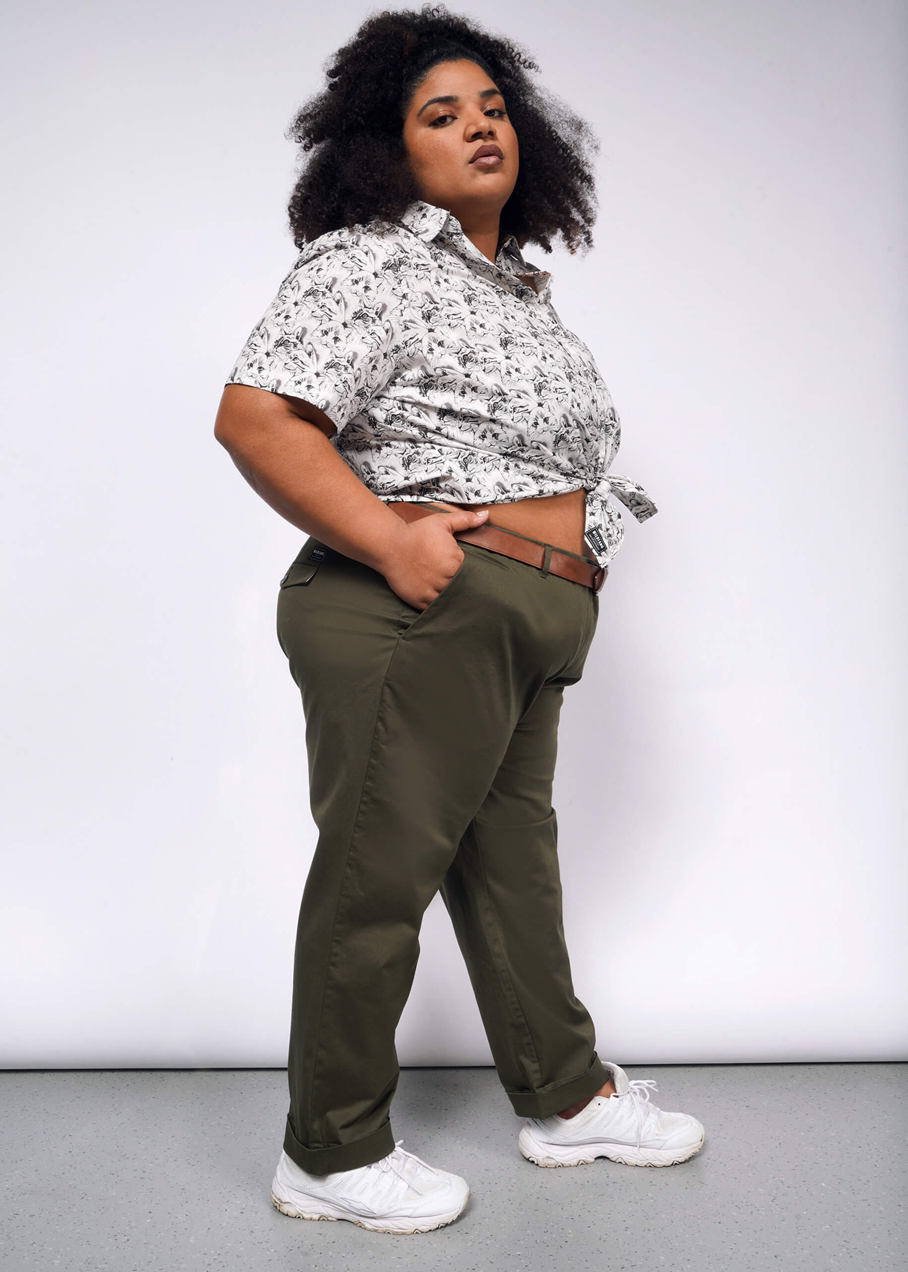 1X=Dressed in The Essential Button Up with Wildfang branded buttons, paired with olive green pants and white sneakers, a person stands confidently against a plain backdrop. With hands in pockets and tailored fit enhancing their look, they gaze directly at the camera.