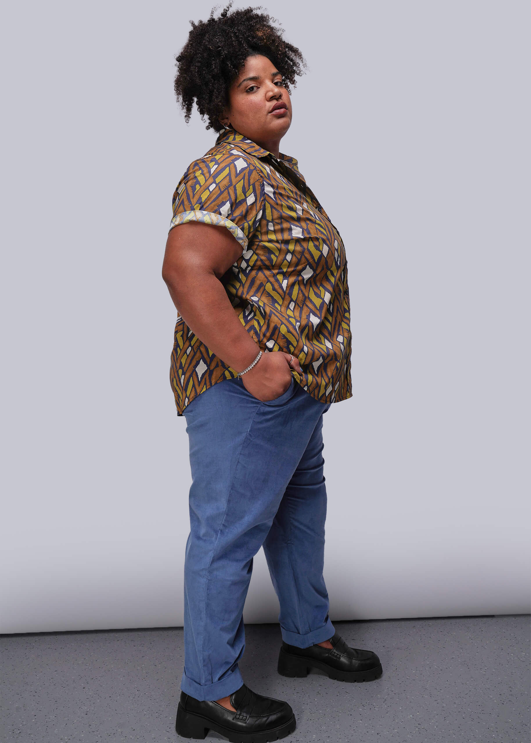 1X=Person wearing a patterned button up shirt and blue pants against a plain background