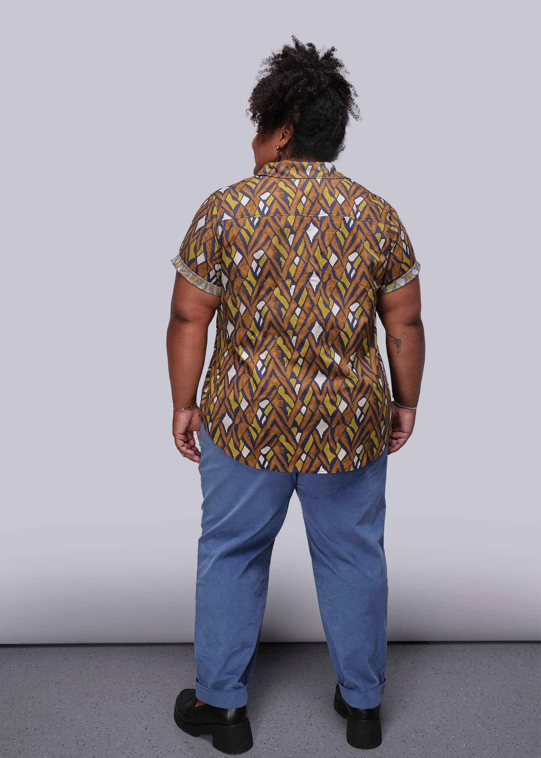1X=Person wearing a patterned shirt and blue pants against a plain background