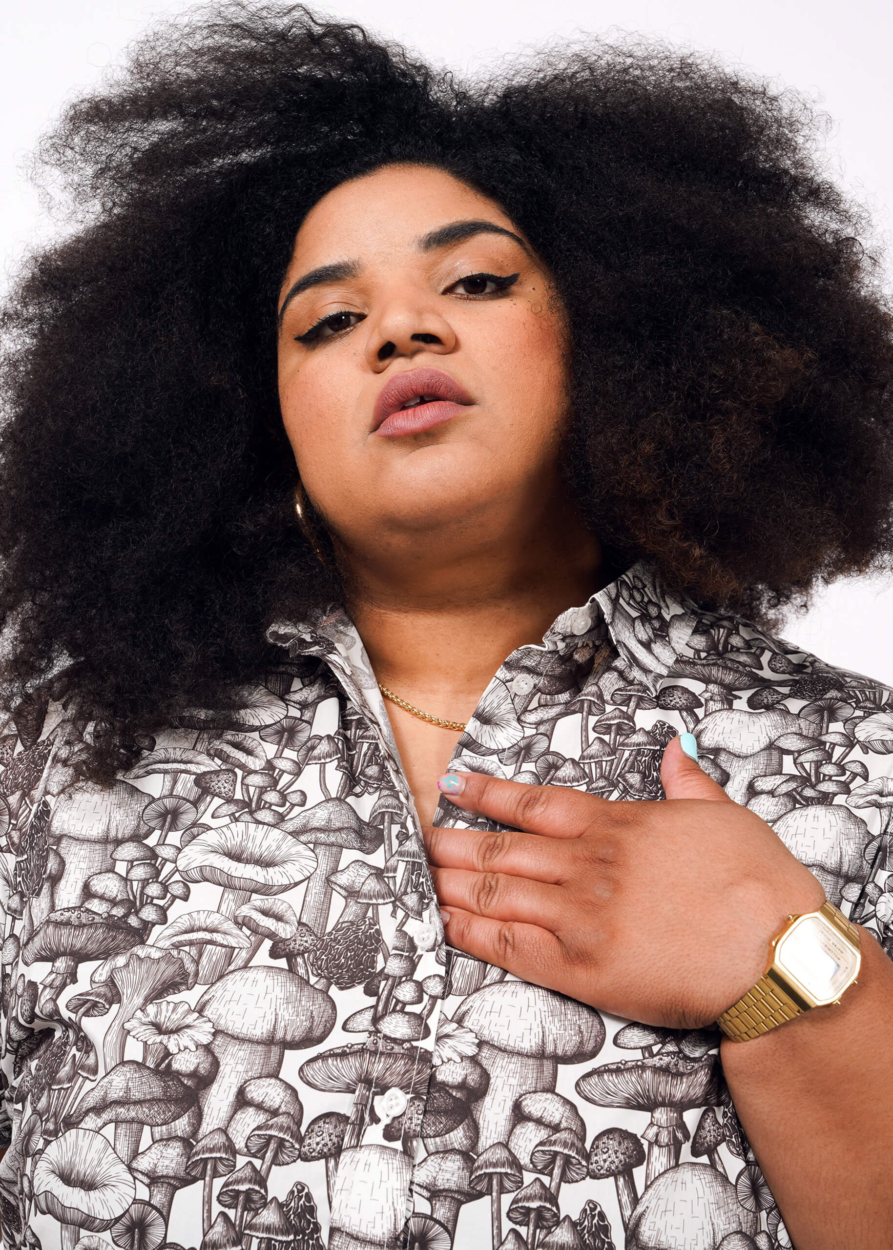 1X=A person with voluminous curly hair wears The Essential Button Up, featuring a mushroom pattern, styled with a gold watch, necklace, and earrings. They look confidently at the camera, one hand on their chest against a plain white background.