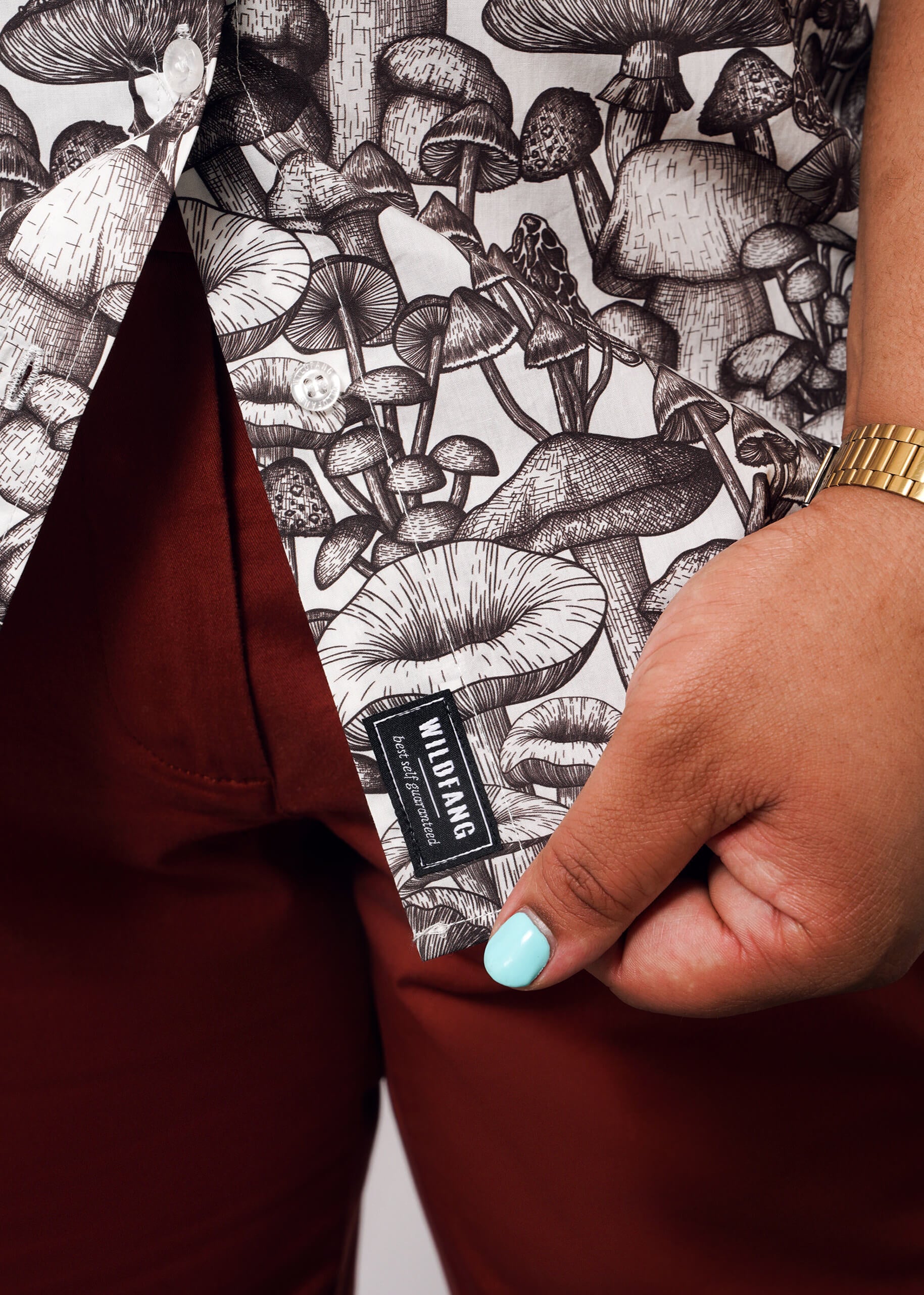 1X=A person with light blue nail polish and a gold watch holds the hem of The Essential Button Up by WILDFANG, a tailored, breathable cotton shirt with a black-and-white mushroom print, paired with reddish-brown pants.