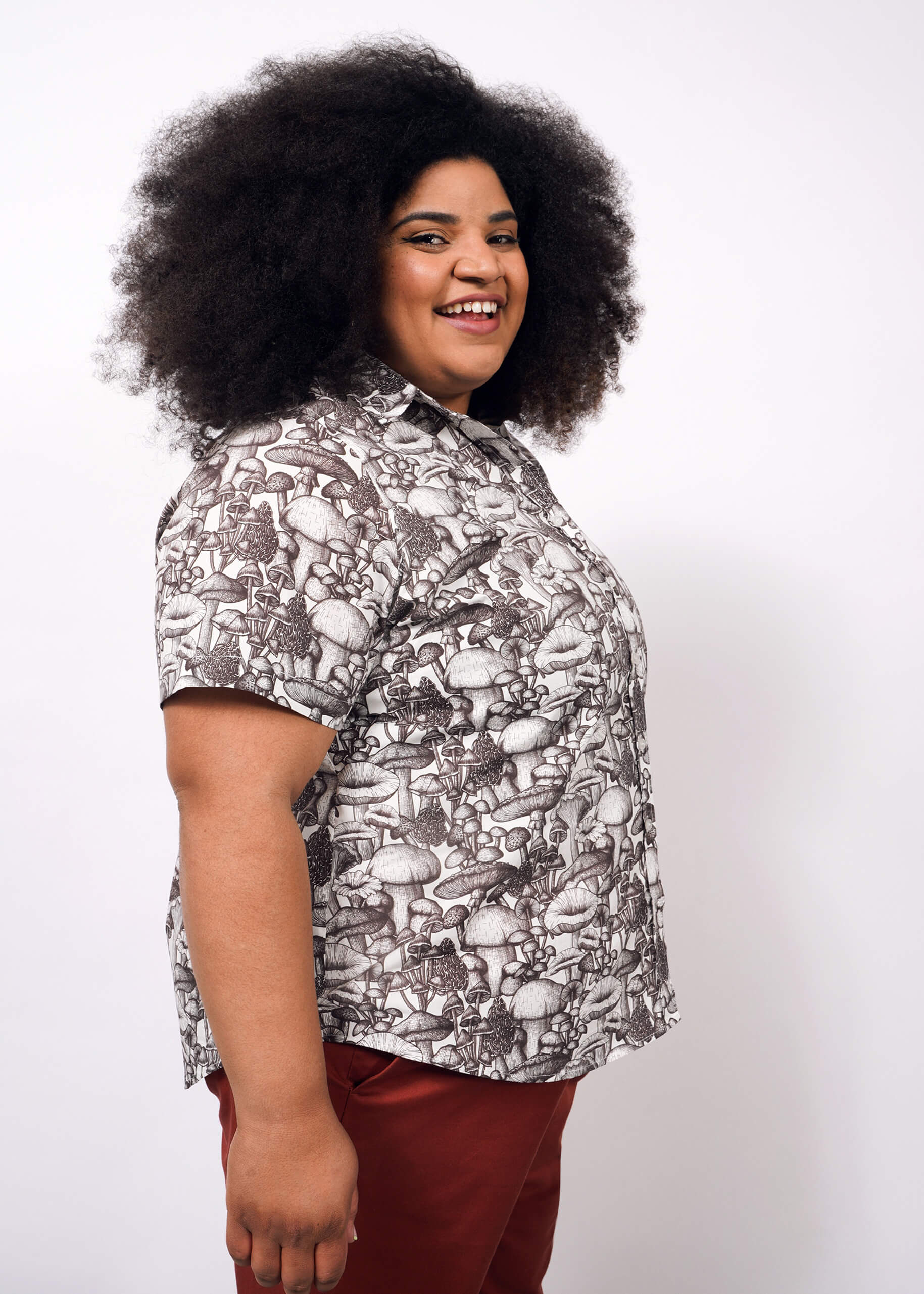 1X= A person with voluminous curly hair smiles sideways, wearing The Essential Button Up—a short-sleeved, breathable cotton shirt with a mushroom pattern and Wildfang-branded buttons—paired with reddish-brown pants against a plain white background.
