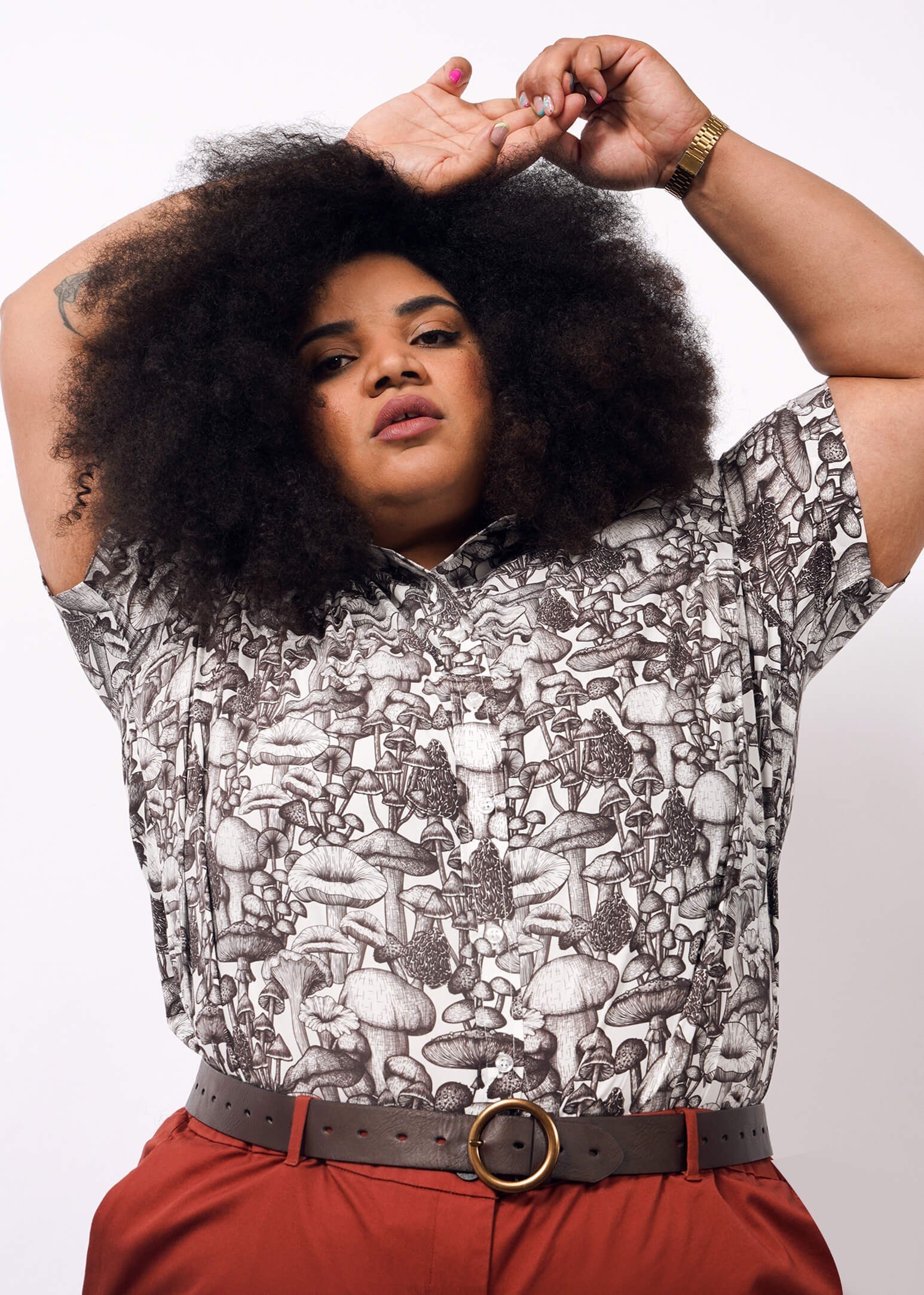 1X=A person with voluminous curly hair poses with arms raised, wearing The Essential Button Up—a patterned mushroom shirt made from breathable cotton with Wildfang branded buttons—along with red pants and a brown belt. Plain white background.