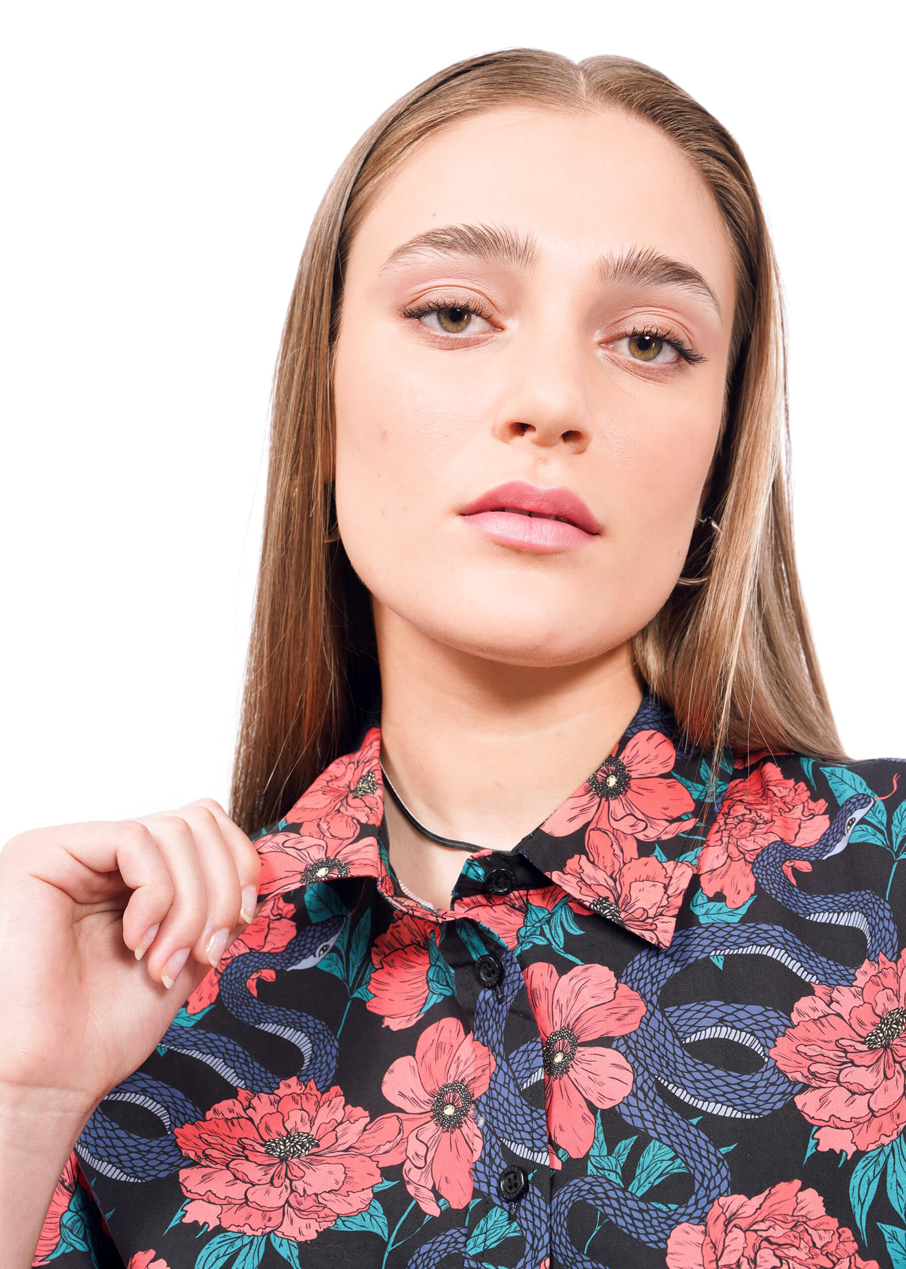 S=A young woman with straight, light brown hair wears The Essential Button Up, featuring pink floral and blue snake patterns. She holds the collar with one hand and looks confidently at the camera against a white background.