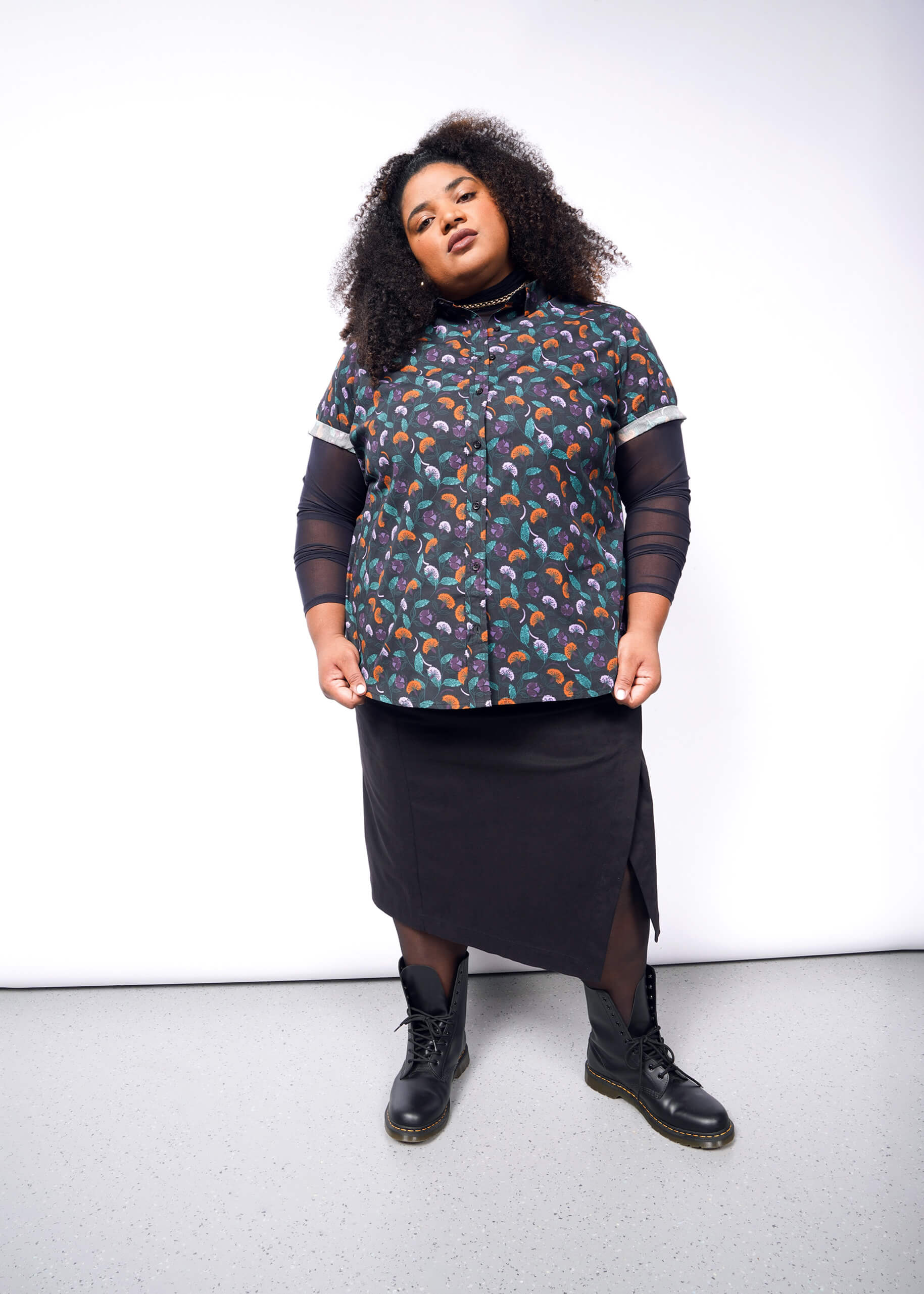 1X= A person with curly hair stands confidently against a white background, wearing The Essential Button Up featuring Wildfang branded buttons. Over a dark long-sleeve top, a black skirt, and black boots complete the tailored fit ensemble. The pose is relaxed yet self-assured.