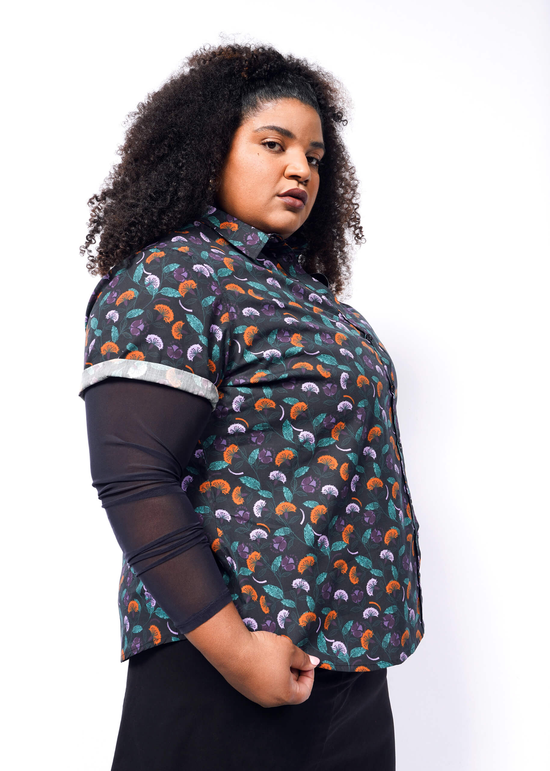1X= A person with curly hair confidently poses against a plain background, wearing The Essential Button Up from Wildfang. This colorful, patterned shirt, made from breathable cotton for comfort, features branded buttons, rolled-up sleeves, and a tailored fit revealing a sheer black top underneath.