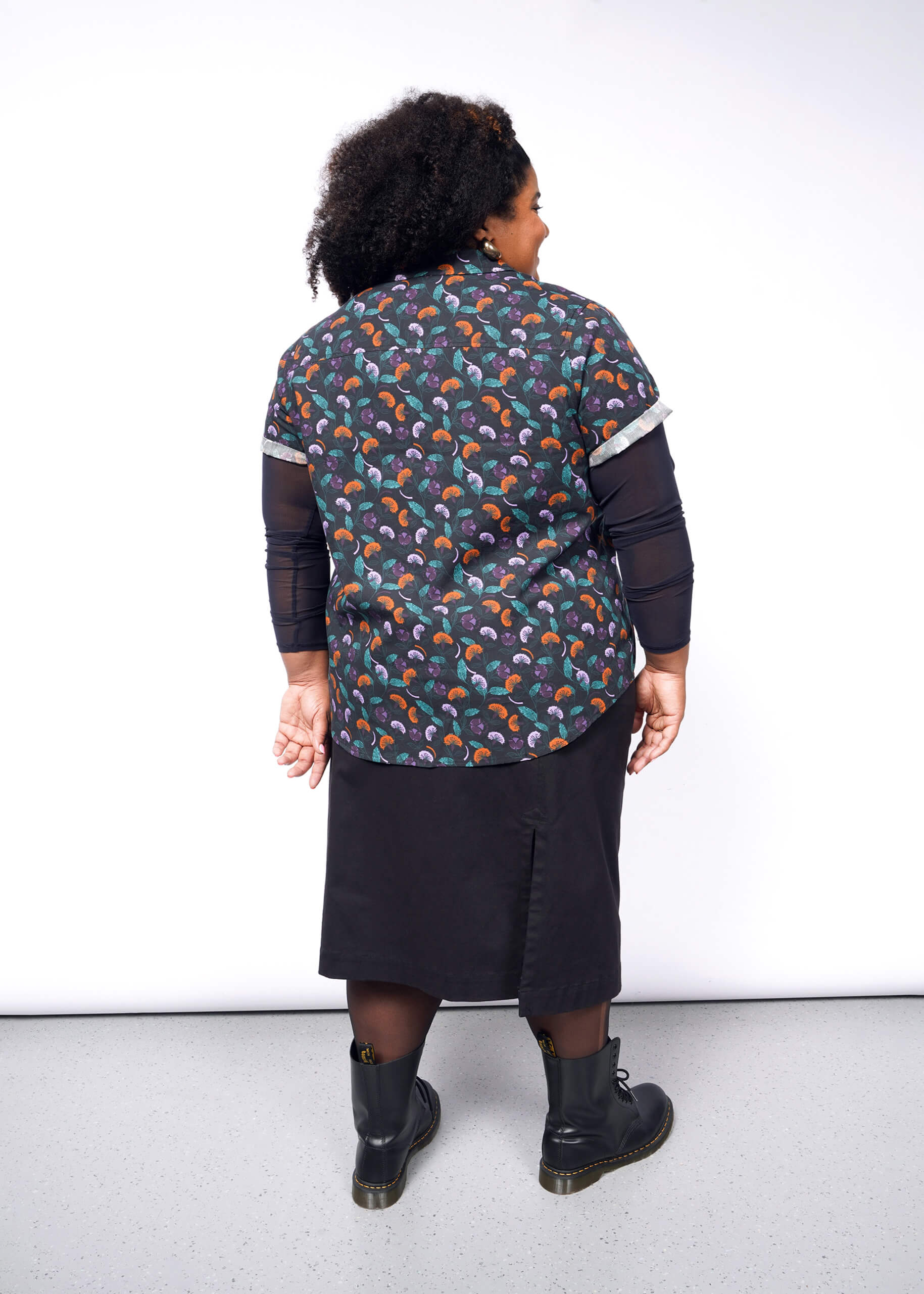 1X=A person with curly hair stands against a white wall, wearing The Essential Button Up, a colorful patterned, breathable cotton shirt. They pair it with a black skirt and boots, arms relaxed at their sides. The background is simple white with gray flooring.