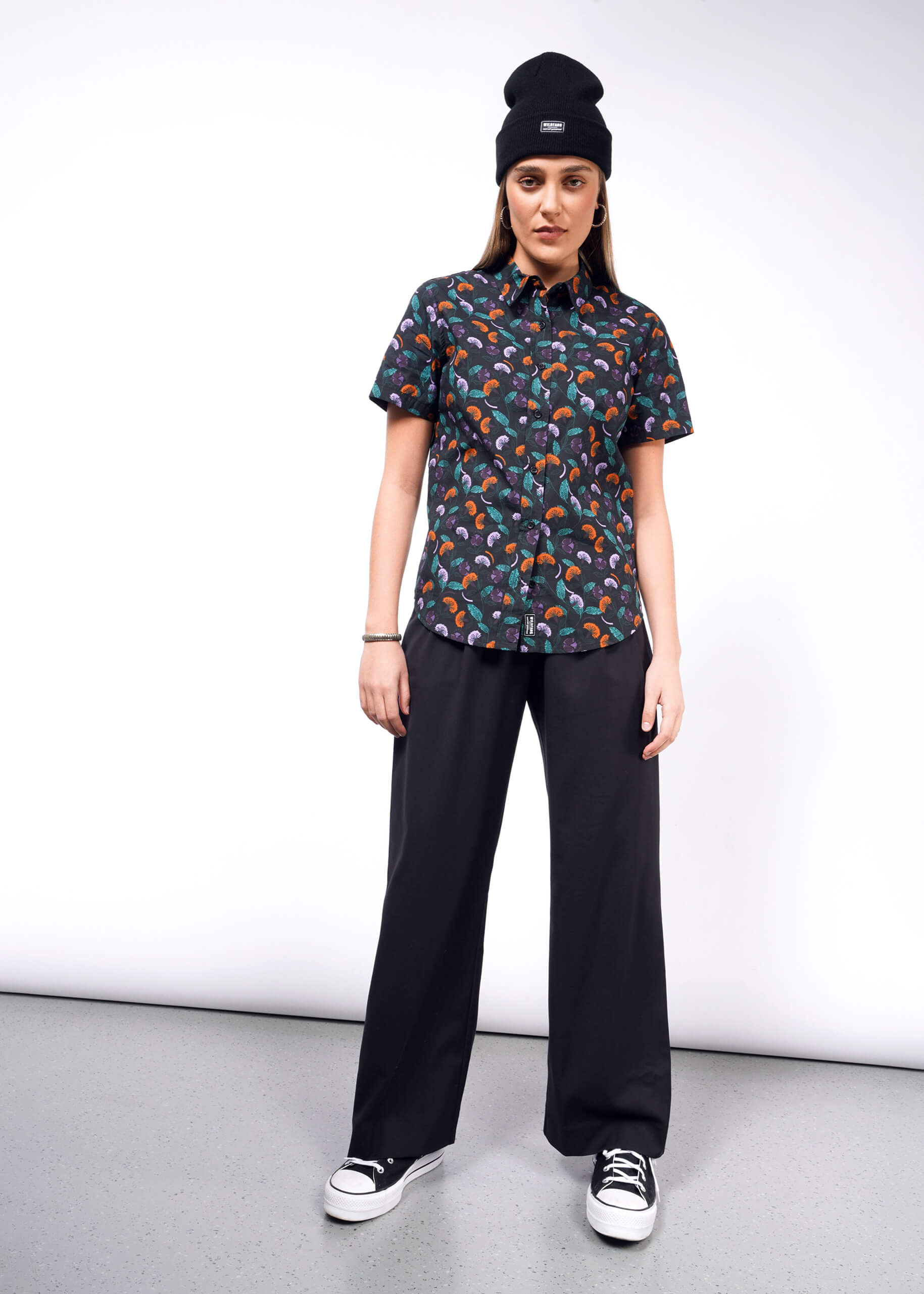 S=Wearing The Essential Button Up, a colorful patterned short-sleeve shirt with Wildfang branded buttons and a tailored fit, they confidently pair it with black wide-legged pants, a black beanie, and sneakers. Standing against a plain white background, they look straight ahead with hands at their sides.
