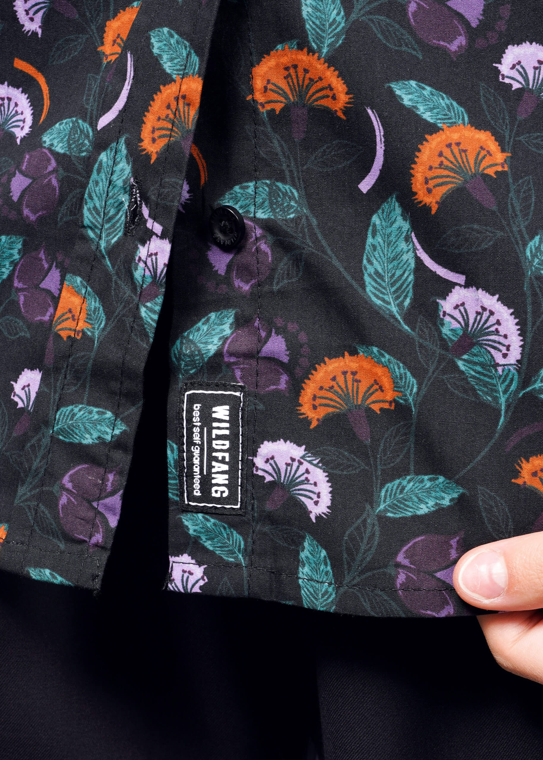 S=Close-up of The Essential Button Up, a black shirt with orange, purple, and green floral patterns. A hand shows off this breathable cotton Wildfang classic with a WILDFANGS label and tailored fit, set against a dark background.