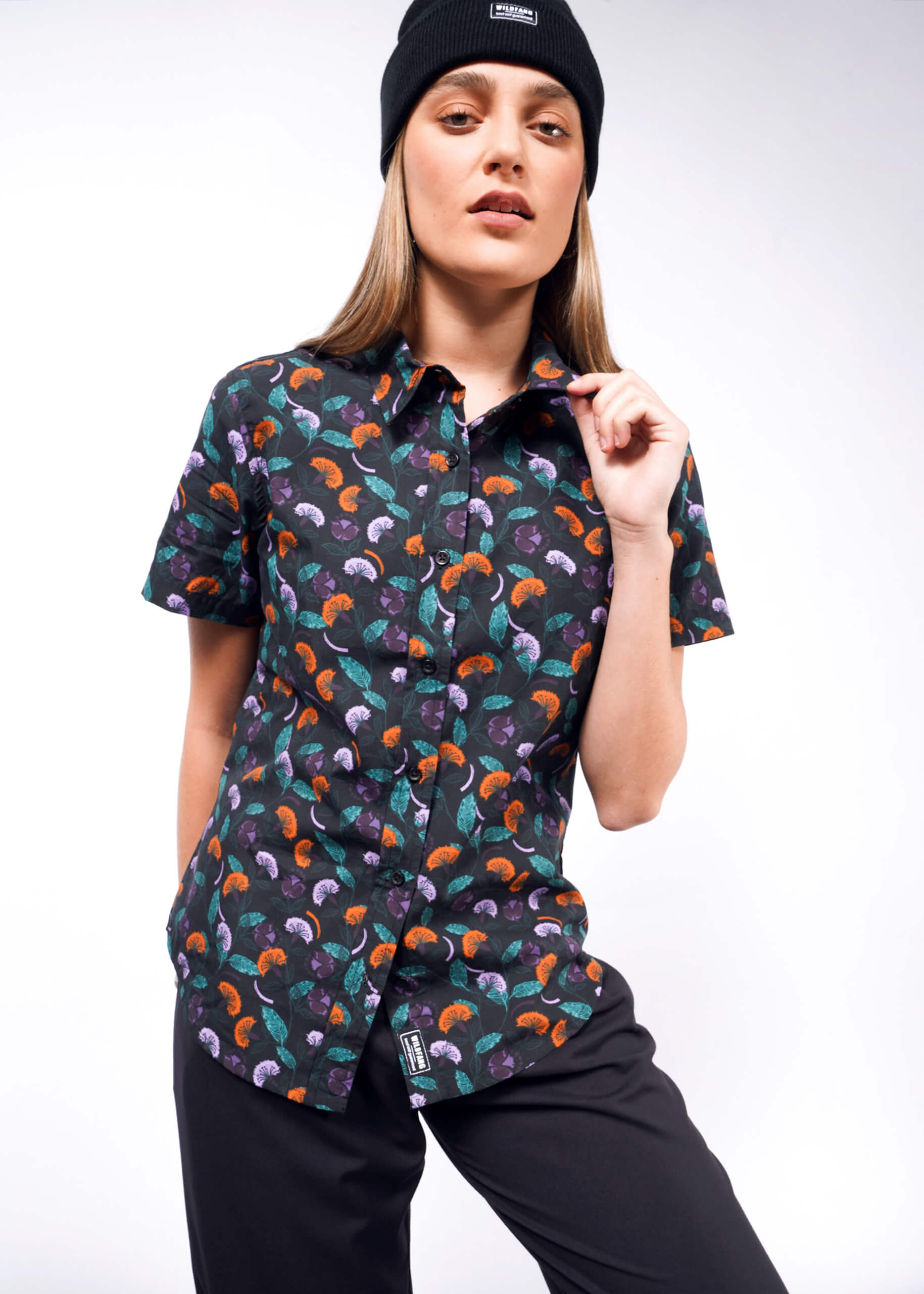 S= A person wearing a black beanie and The Essential Button Up, a short-sleeve shirt featuring a colorful mushroom pattern in breathable cotton, pairs it with tailored fit black pants while posing casually against a white backdrop.