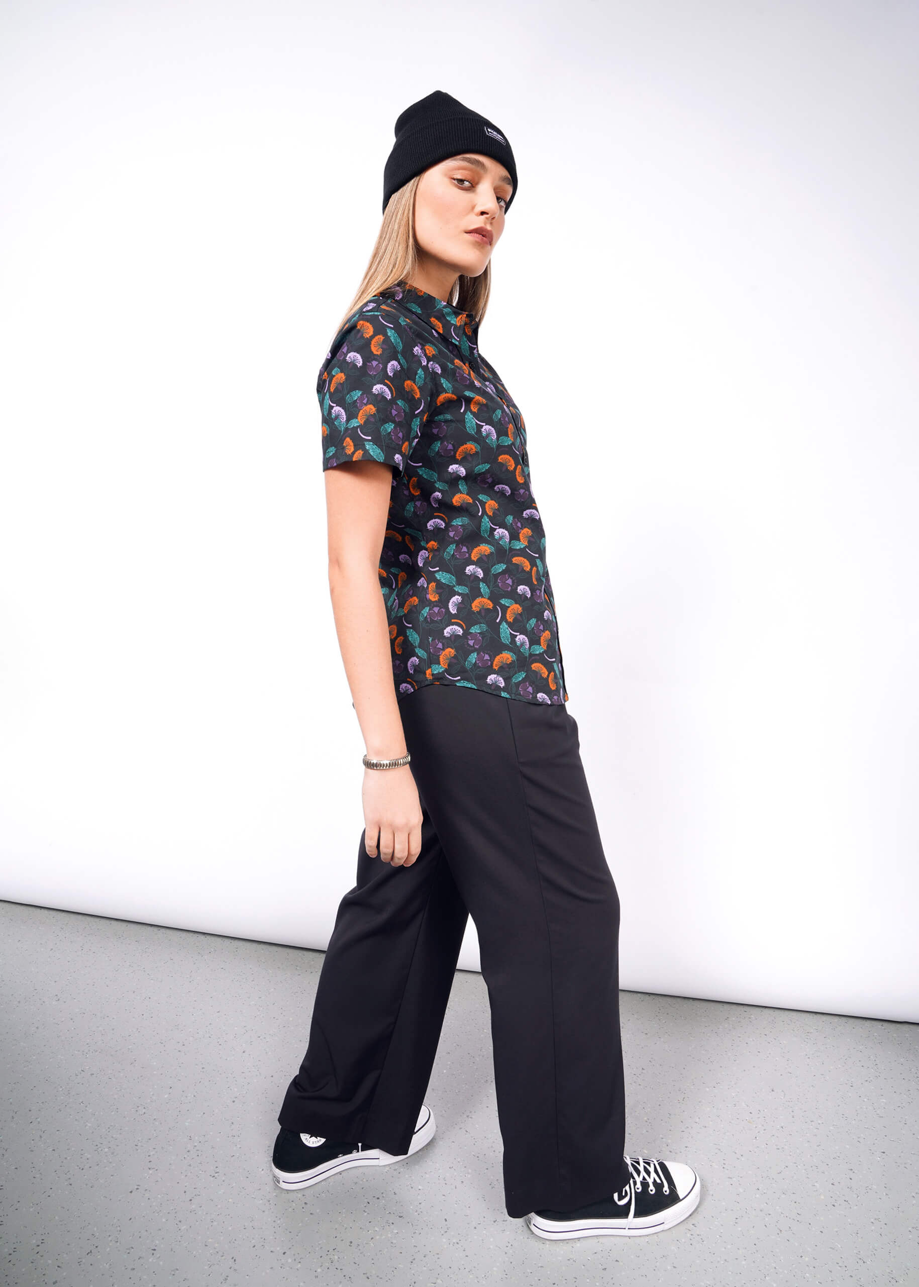 S=A person confidently wears a black Essential Button Up by Wildfang in breathable cotton with a colorful pattern, complemented by black wide-leg pants, shoes, and beanie. Theyre looking to the side against a plain white backdrop.