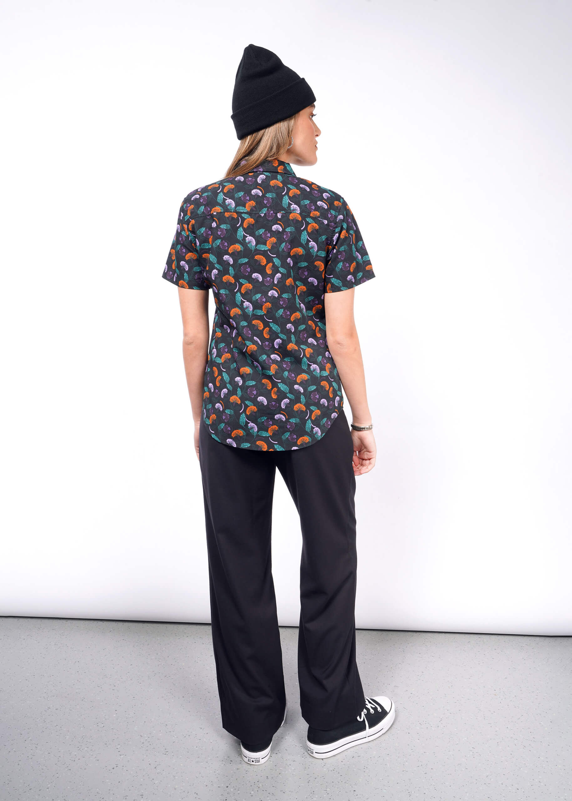 S=A person wearing a black beanie, a short-sleeved Essential Button Up with a colorful abstract pattern, tailored fit black pants, and sneakers stands facing away from the camera against a plain light background.
