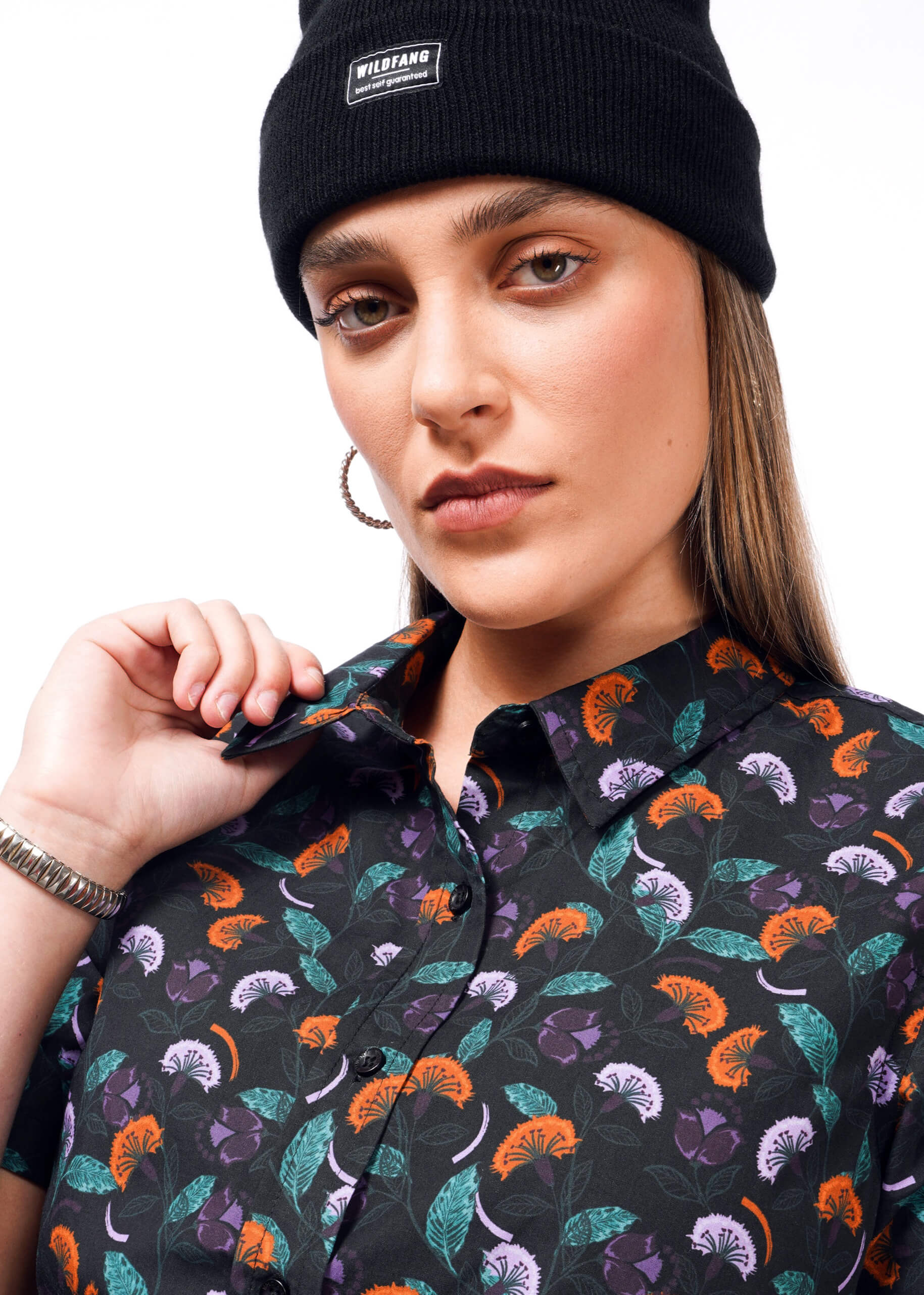 S= A person in a black beanie and hoop earrings looks at the camera with a neutral expression. They wear The Essential Button Up by Wildfang, featuring a vibrant floral pattern in purple, orange, and green on black, offering breathable cotton comfort.