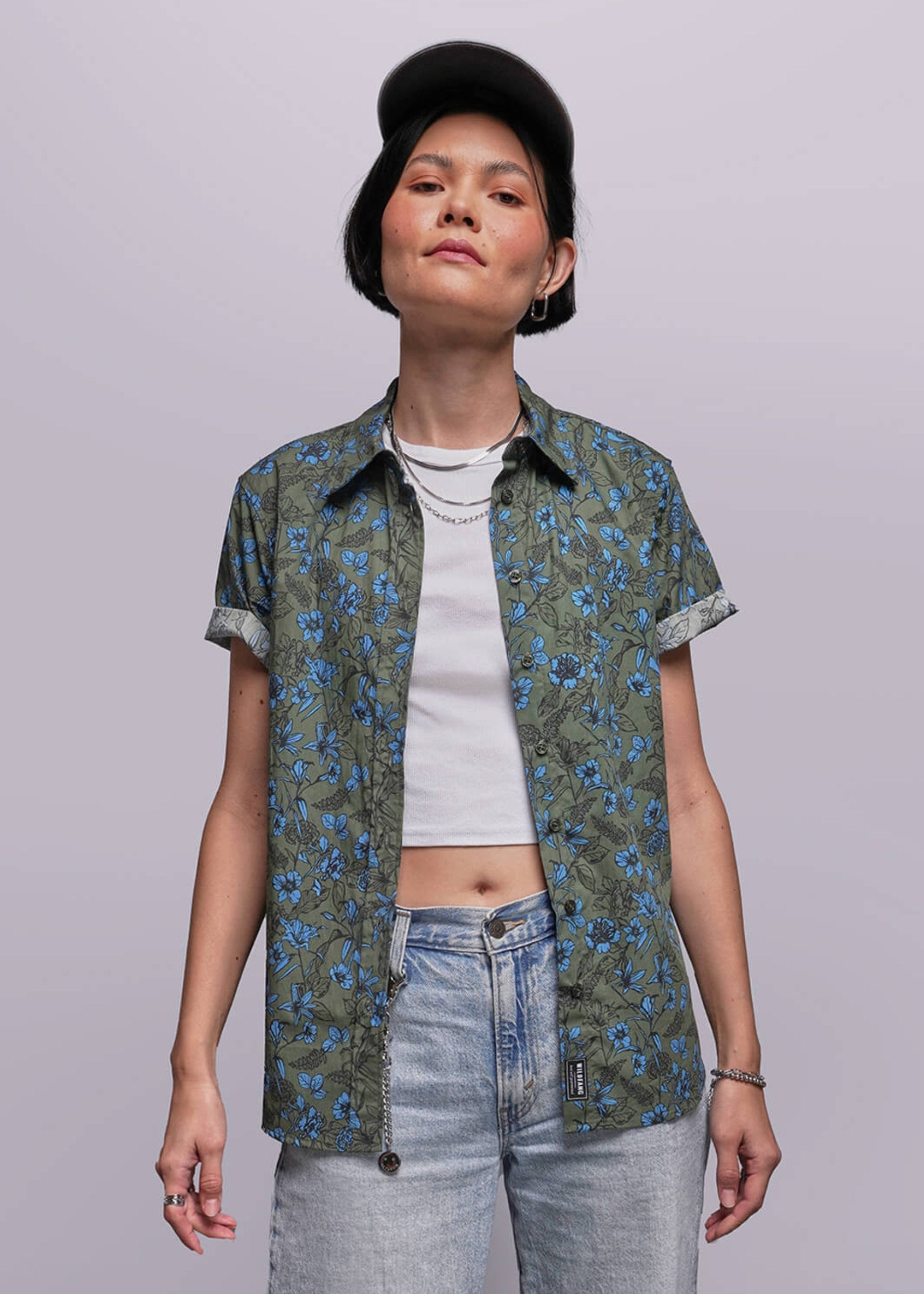 S=Person wearing a floral short-sleeve shirt over a white crop top with blue jeans on a plain background