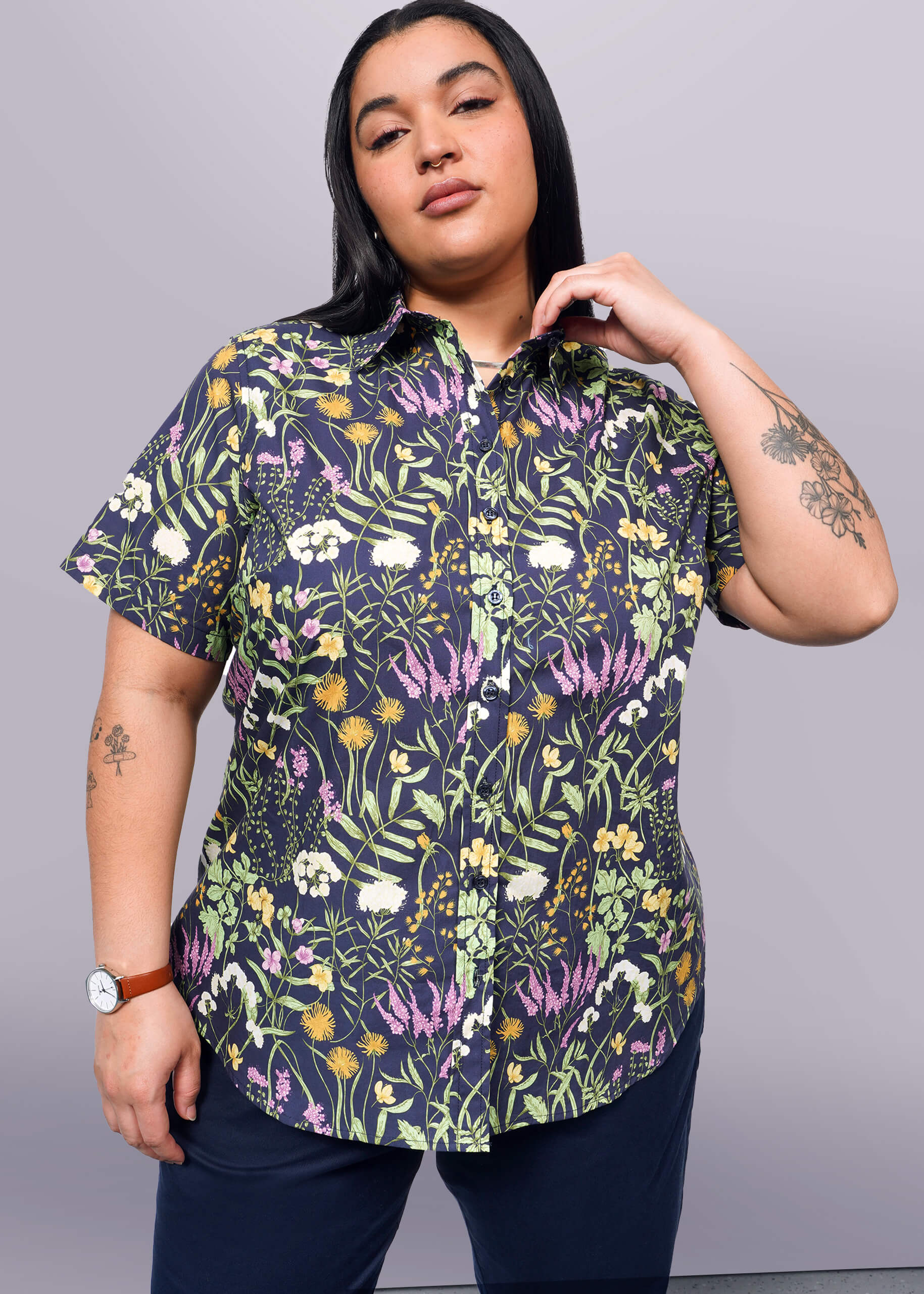 1X= Person with long black hair looks at camera while holding collar of short sleeve button up shirt with botanical print.
